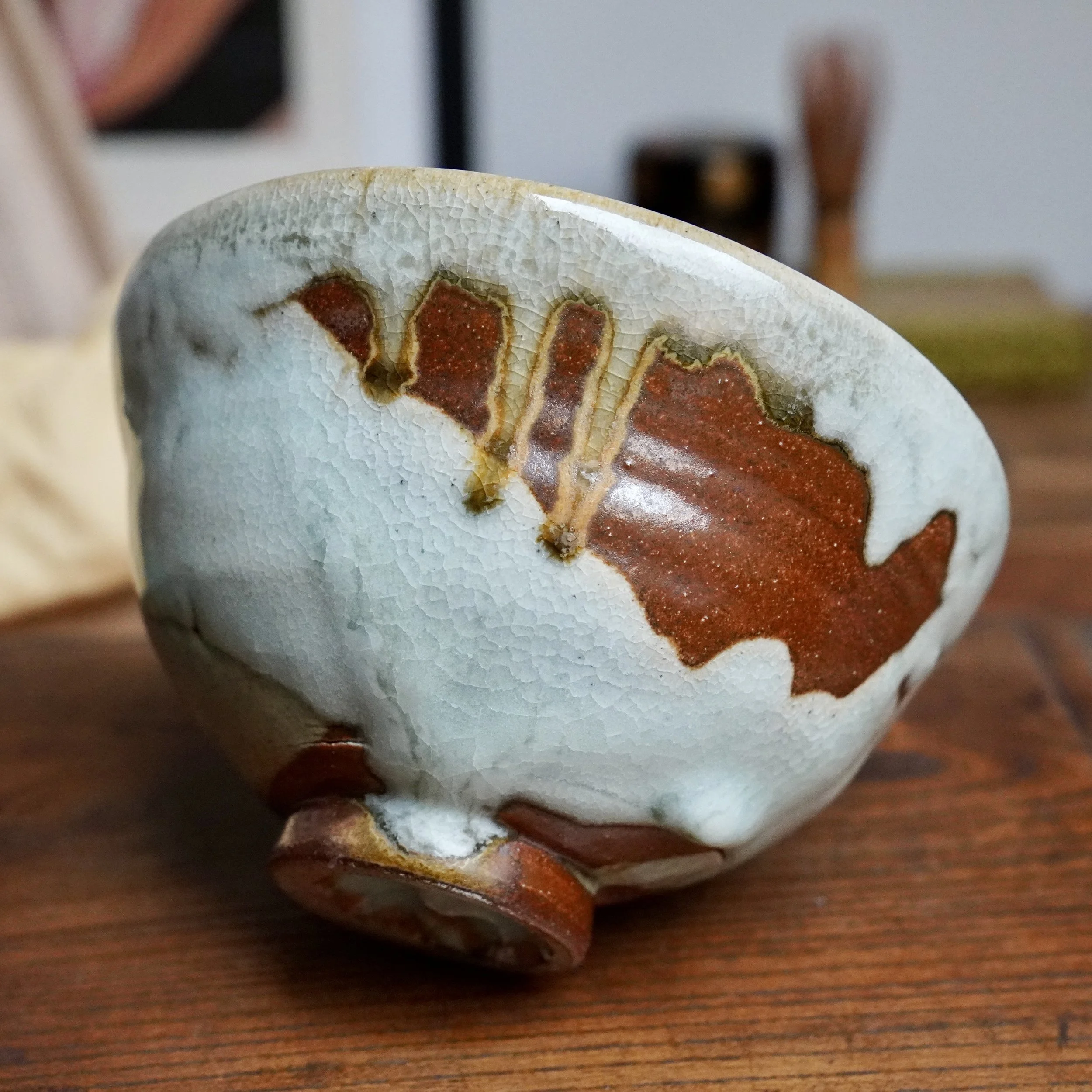 Chawan showing pale, crackled blue-grey glaze flowing into rustic red-brown glaze with ochre streaks. Unglazed foot is earthy red-brown.