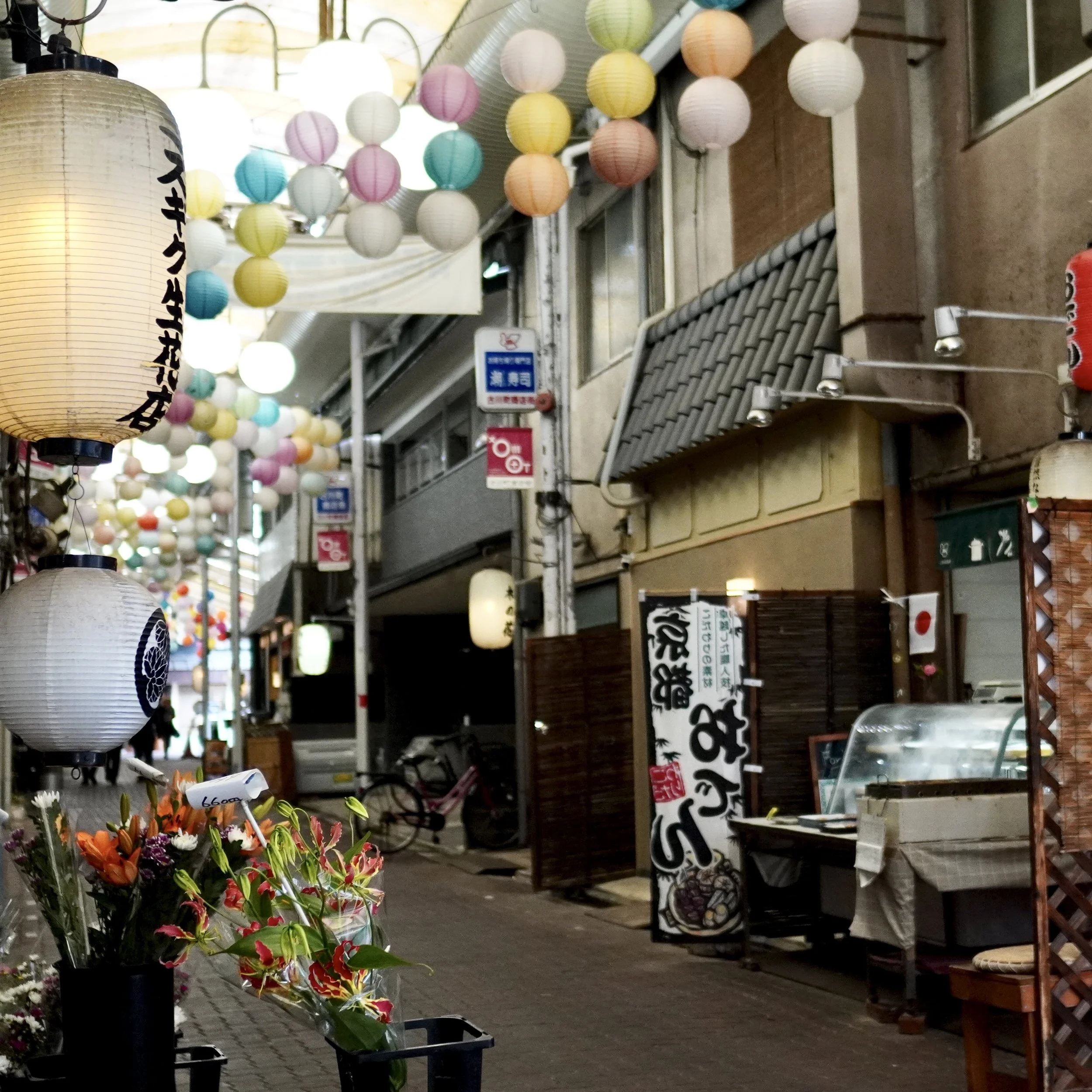 A narrow covered shopping street in Kyoto, known as Teramachi-dori shotengai, decorated with colorful hanging lanterns and traditional shop fronts selling local flowers and Japanese goods.