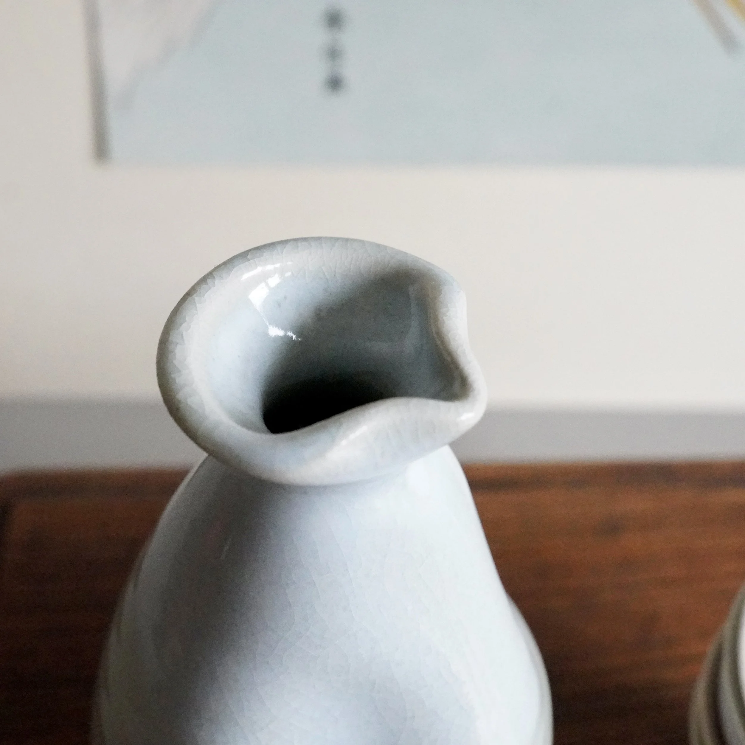Pale blue-grey ceramic sake tokkuri, close-up of its pouring spout and neck. The surface features fine crazing, and the interior is darker. Blurred background with wood and a light wall.