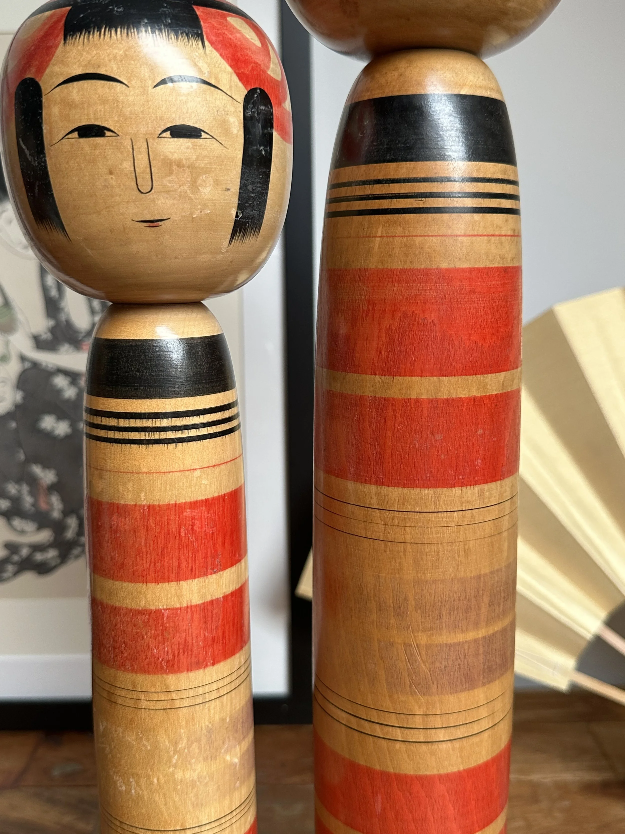 Abe Ichirō Tsuchiyu kokeshi pair, one in the foreground showing its wooden face with black hair and simple features, both featuring bodies banded in red, black, and natural wood.