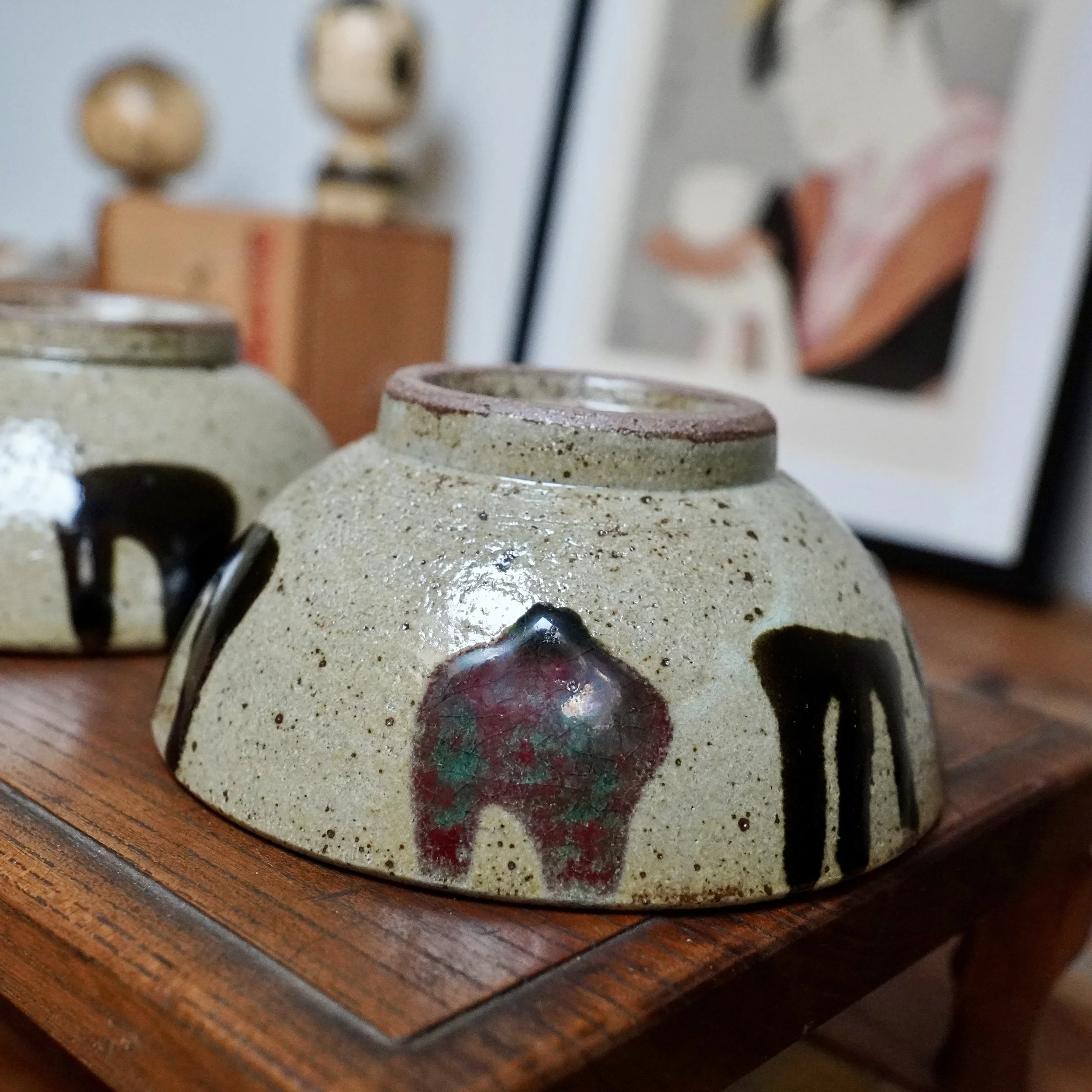 Two Maruta Nobumasa hira-chawan, upside down on wood. Pale ceramic with black glaze drips, one bowl has wine-red and green glazes.