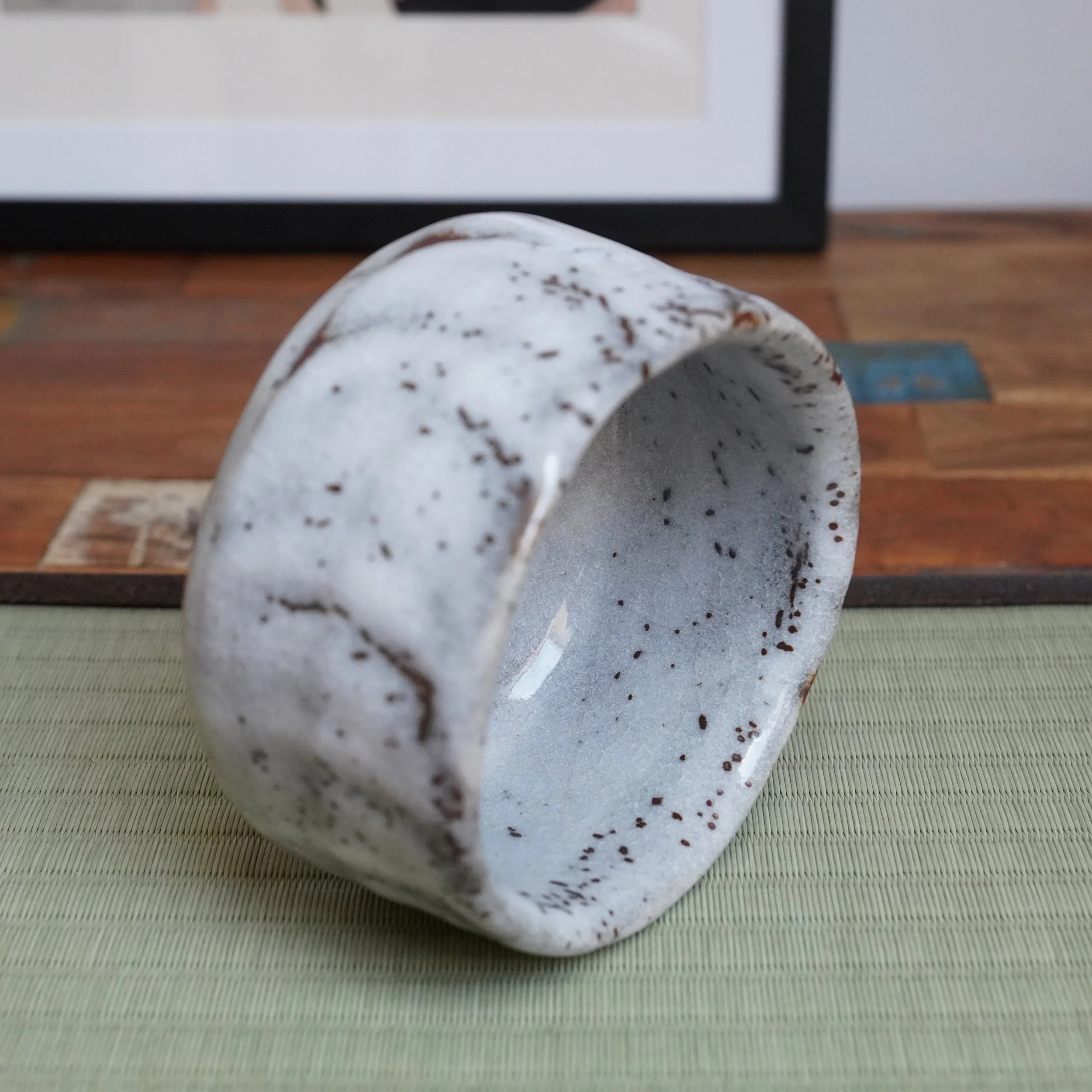 Katō Shōzan's Oni-Shino chawan, tilted on a tatami mat, showing its irregular white body with shallow hollows and rust brown specks, and a crackled interior.