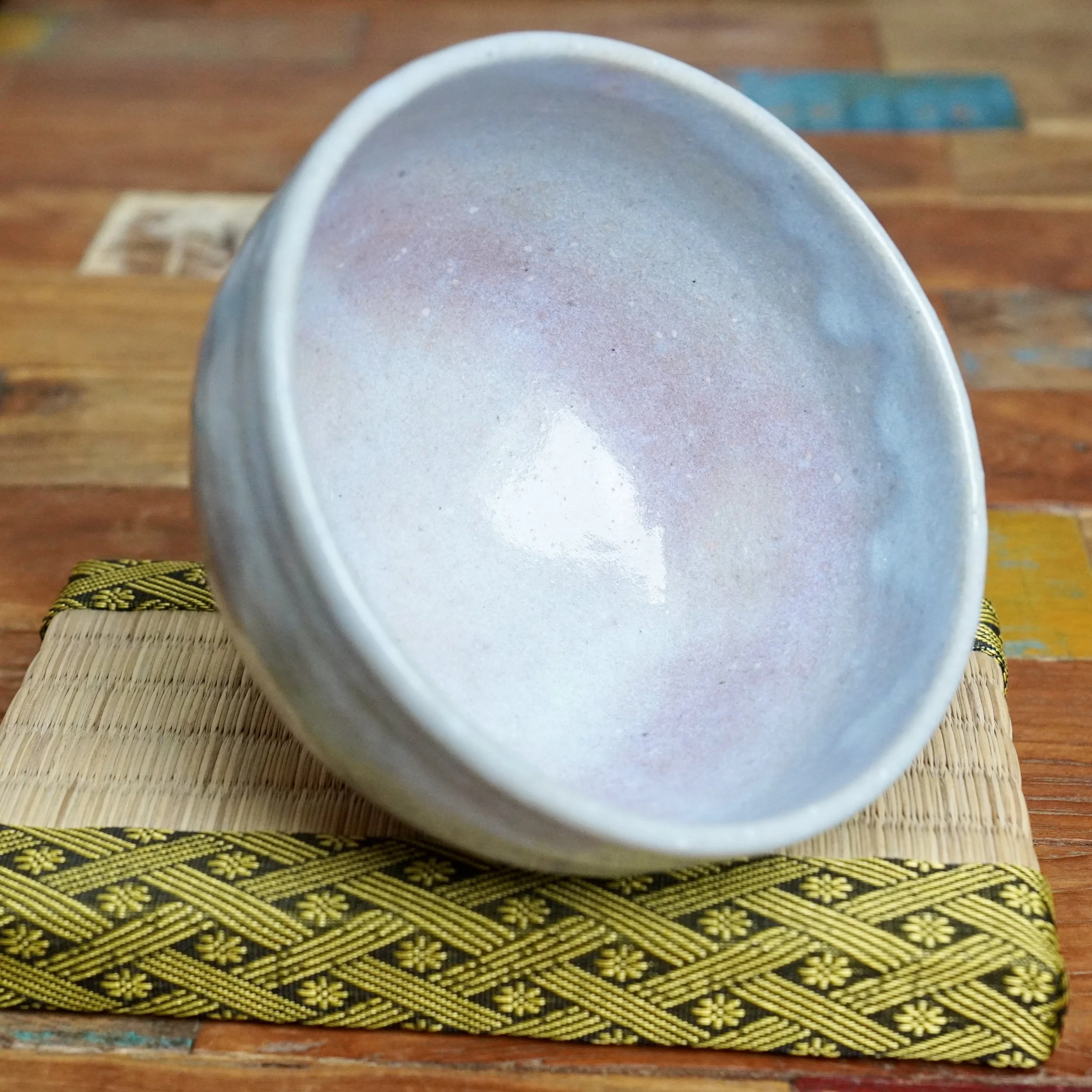 Moriwaki Fumitada chawan, interior view showing soft white, blue-grey, and lilac glaze. It rests on a straw mat with a yellow-gold patterned border.