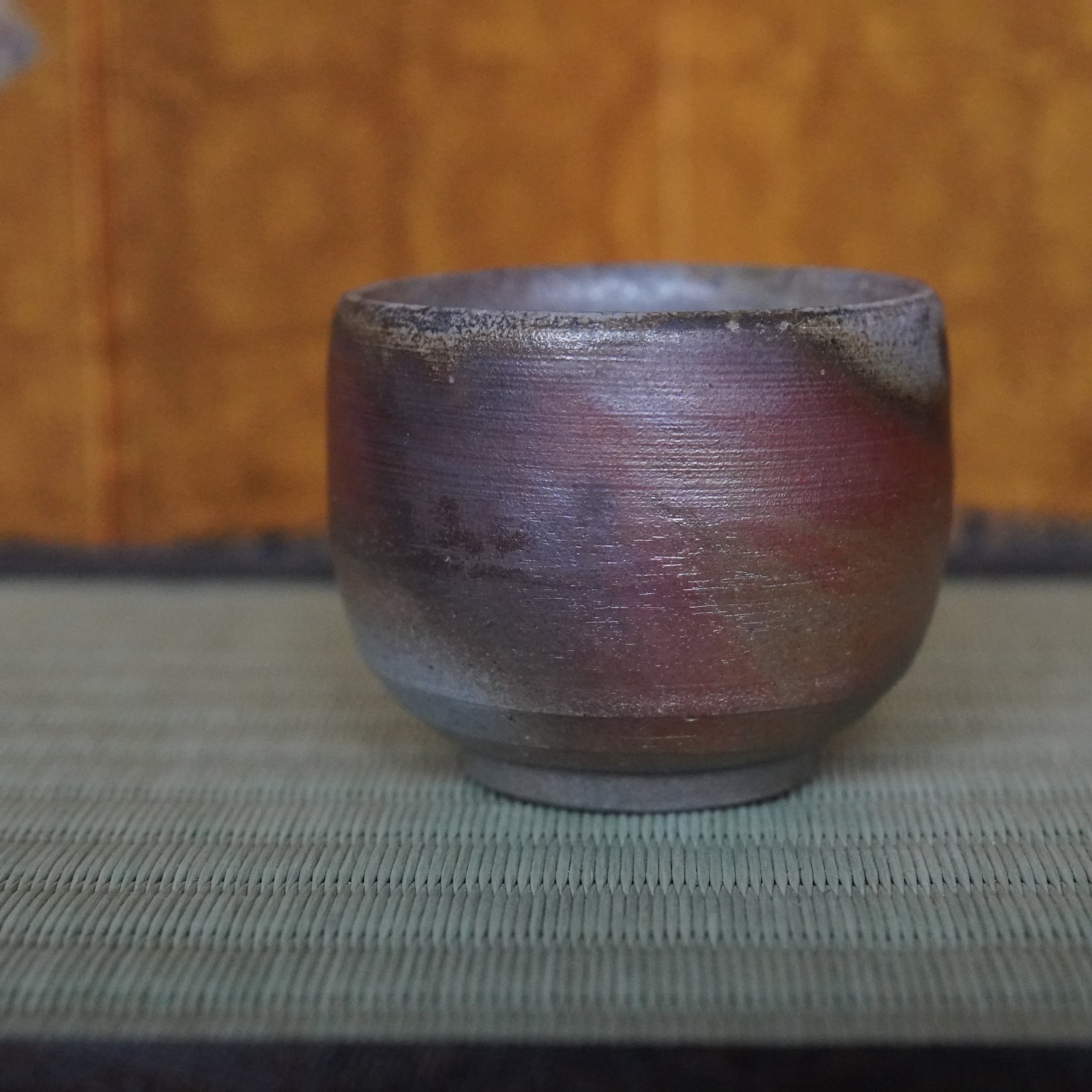 Yamamoto Sōshū Bizen-yaki guinomi, a dark, textured cup with curving reddish hidasuki flame marks, sitting on a woven mat.