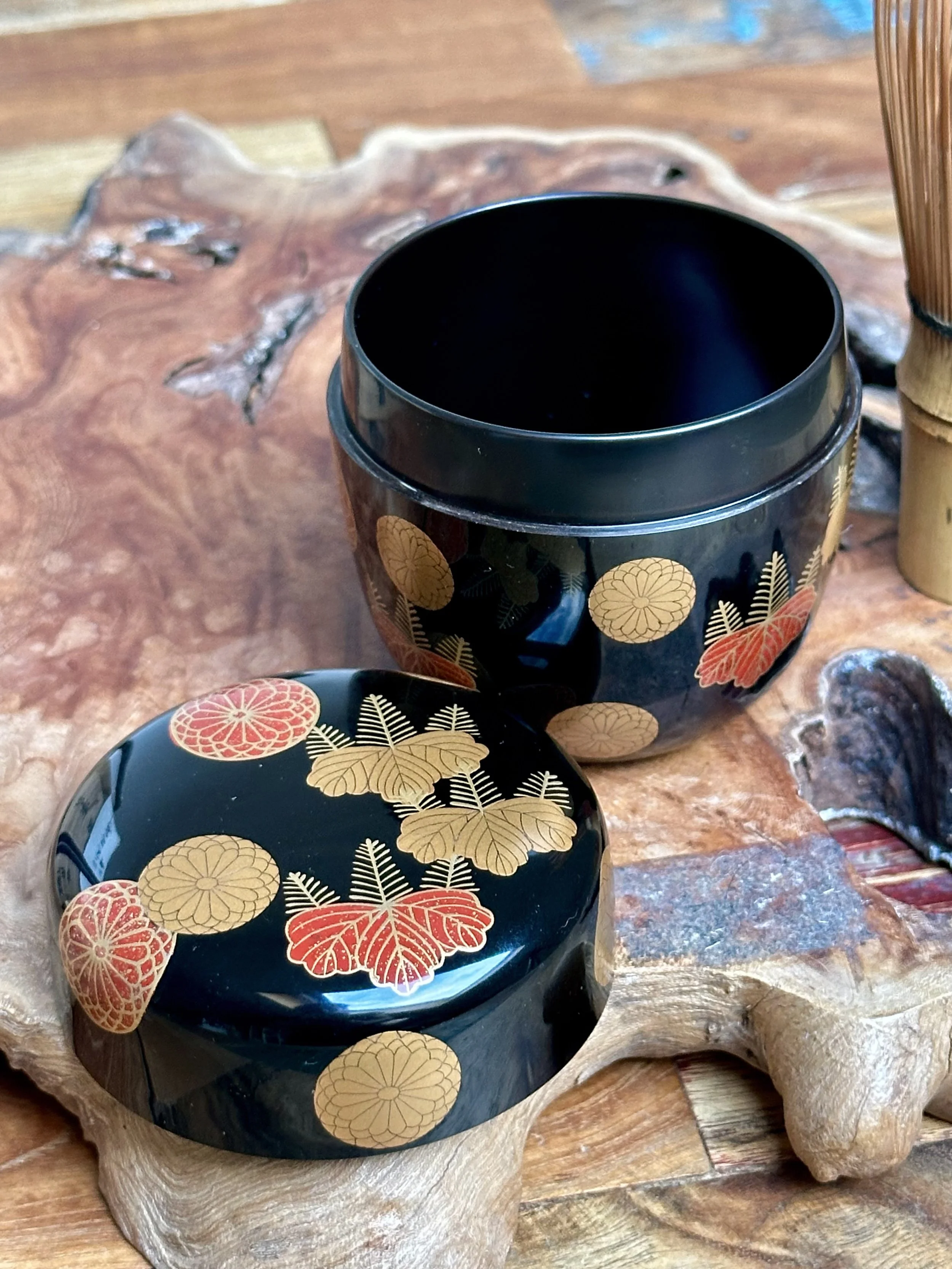 Close-up of a black lacquered natsume tea caddy with its lid off, showing gold and red chrysanthemum and paulownia crests. It rests on textured wood, with a bamboo whisk in the blurred background.