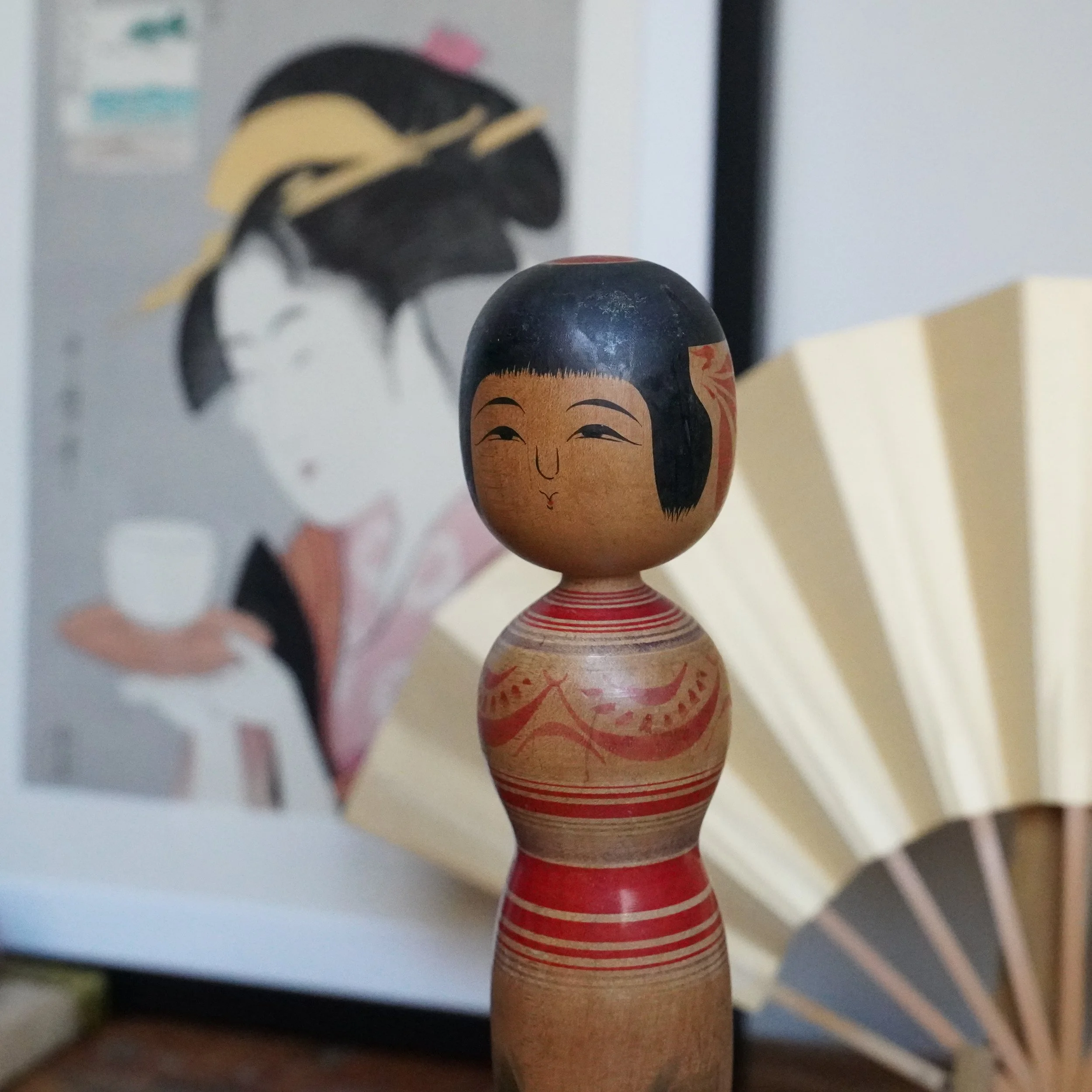 Tsugaru kokeshi by Sasamori Junichi, with black hair, a smiling face, and an hourglass body decorated with red peony and stripes. Blurred background of a Japanese print and fan.