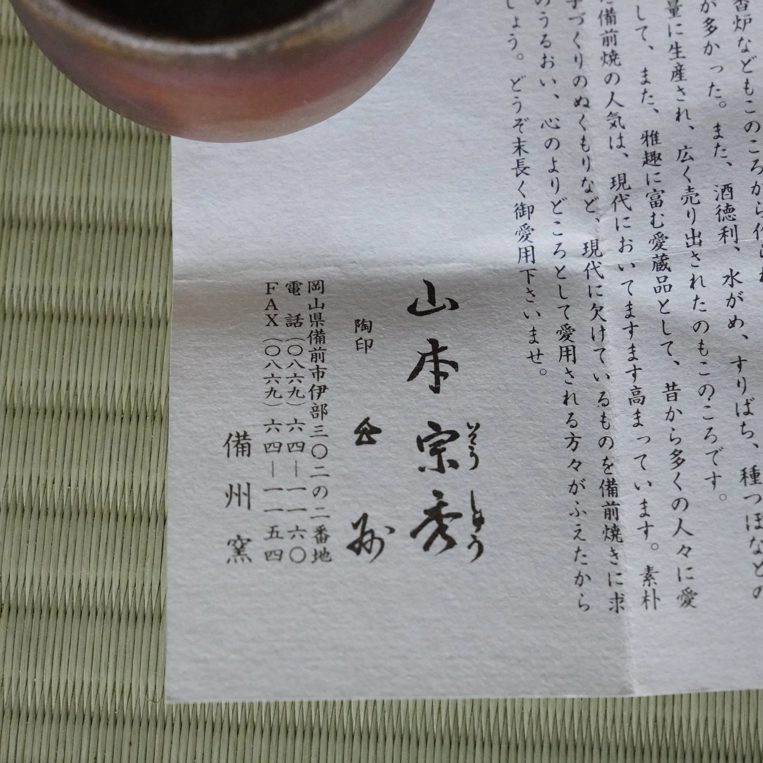 Shiori from Yamamoto Sōshū, showing his calligraphy signature and Bishū-gama kiln details. A reddish-brown Bizen-yaki guinomi is partially visible on a tatami mat.