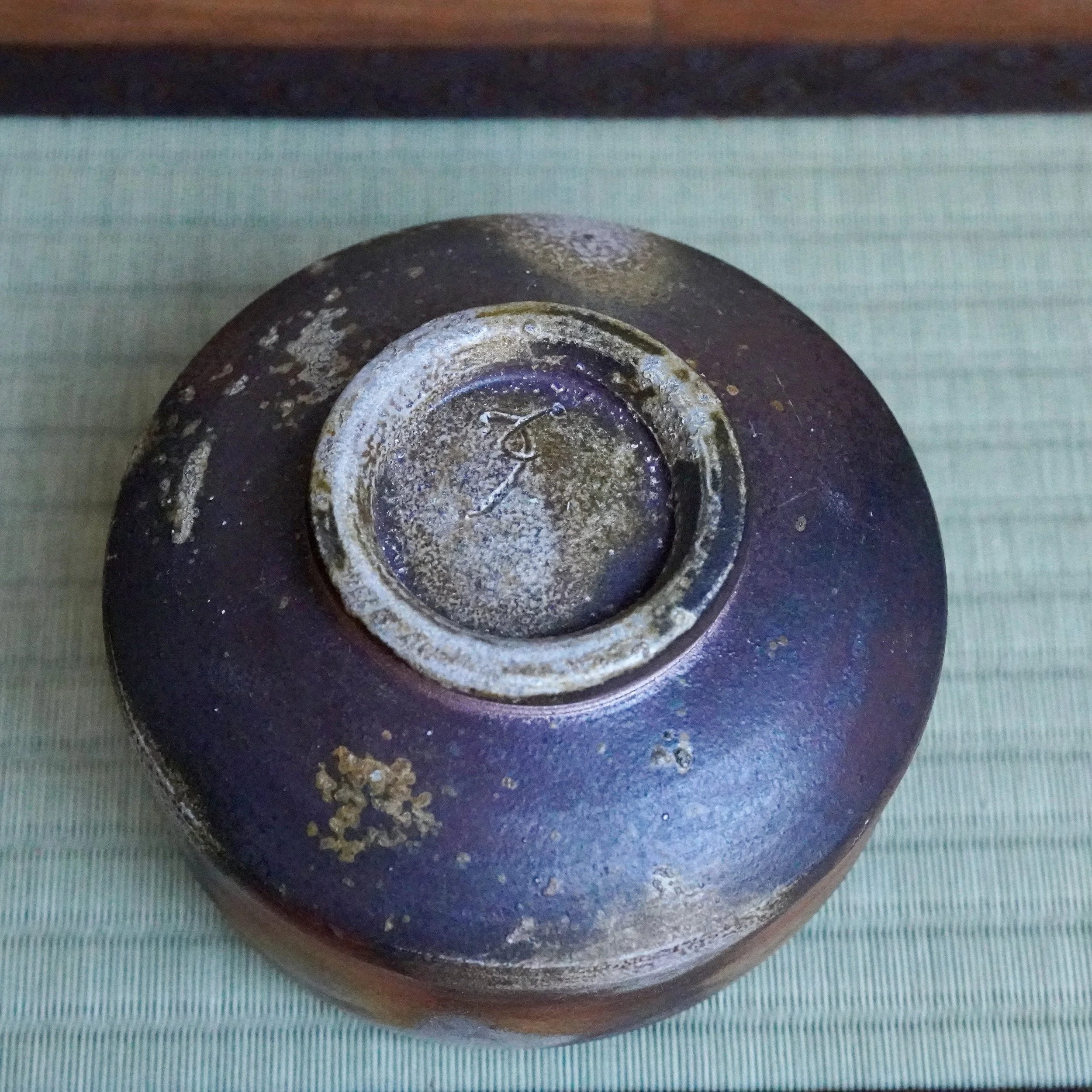 Underside of a Bizen chawan by Inomata Masaaki, showing dark purplish-brown clay, lighter ash deposits, and a distinctive seal inside the footring.