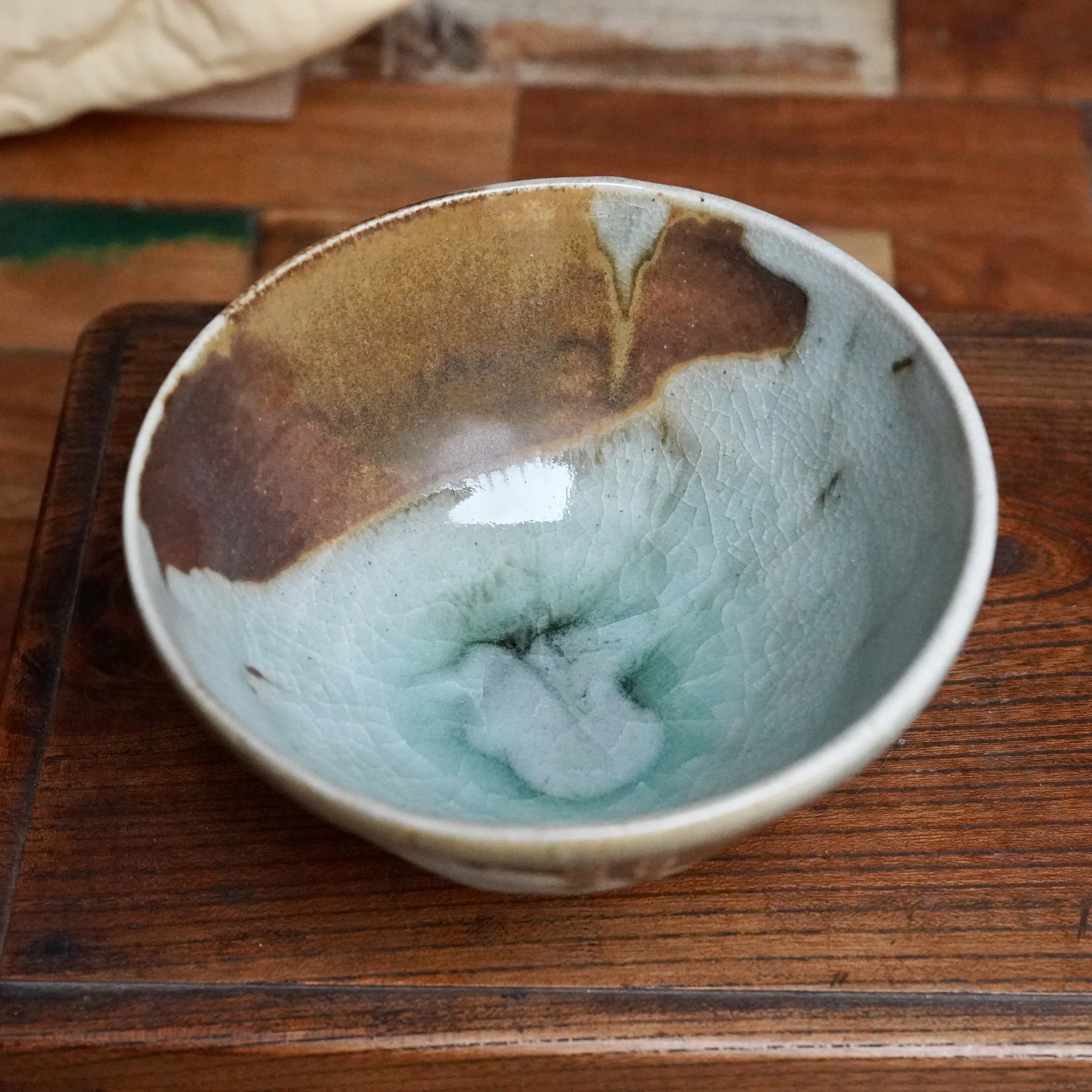 Sansōan chawan with crackled blue-green interior glaze, flowing rust-brown and ochre glaze on one side, presented on a wooden surface.