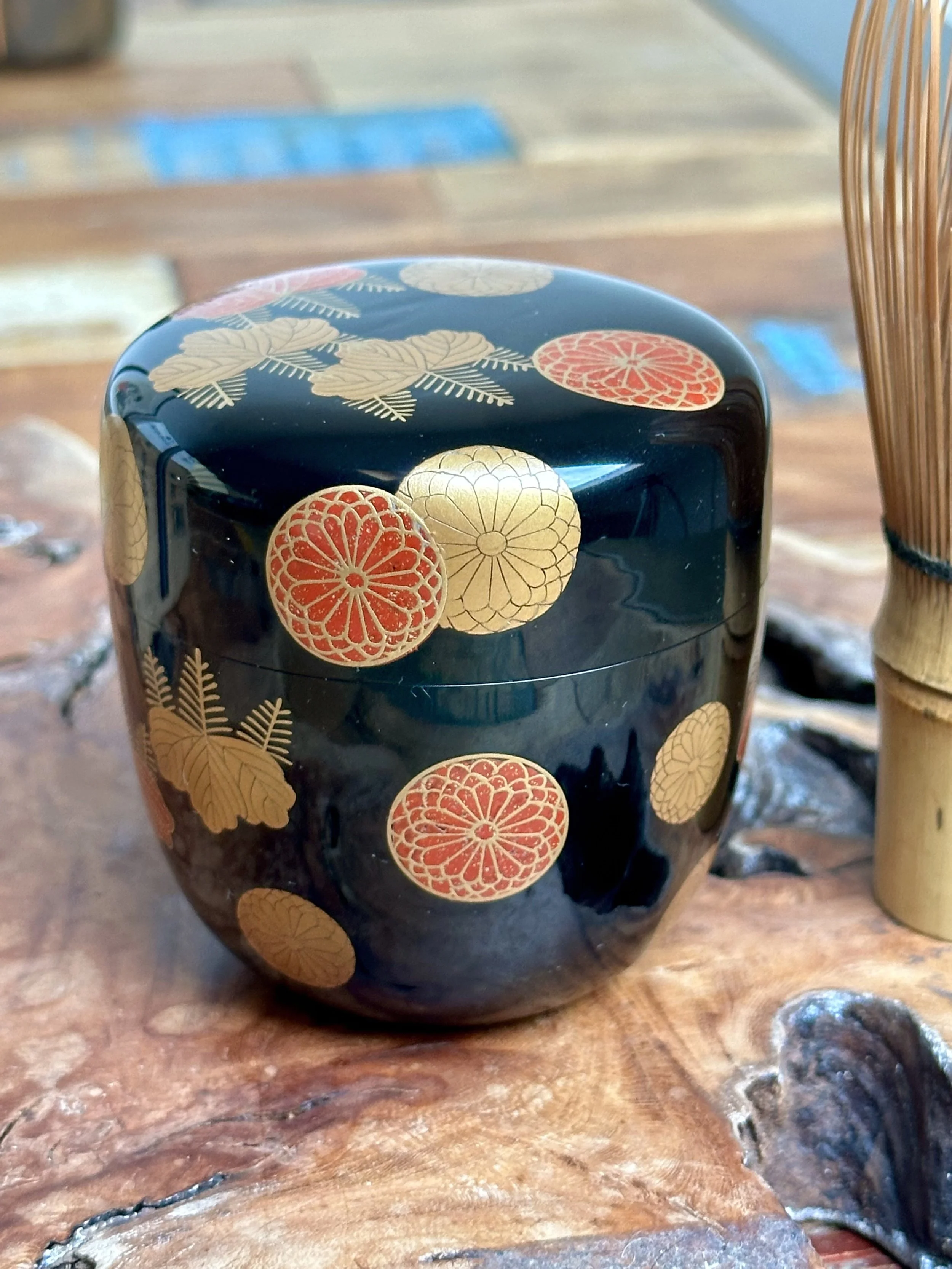 Glossy black makie natsume tea caddy with scattered gold paulownia and red chrysanthemum crests, on a wooden surface next to a bamboo whisk.