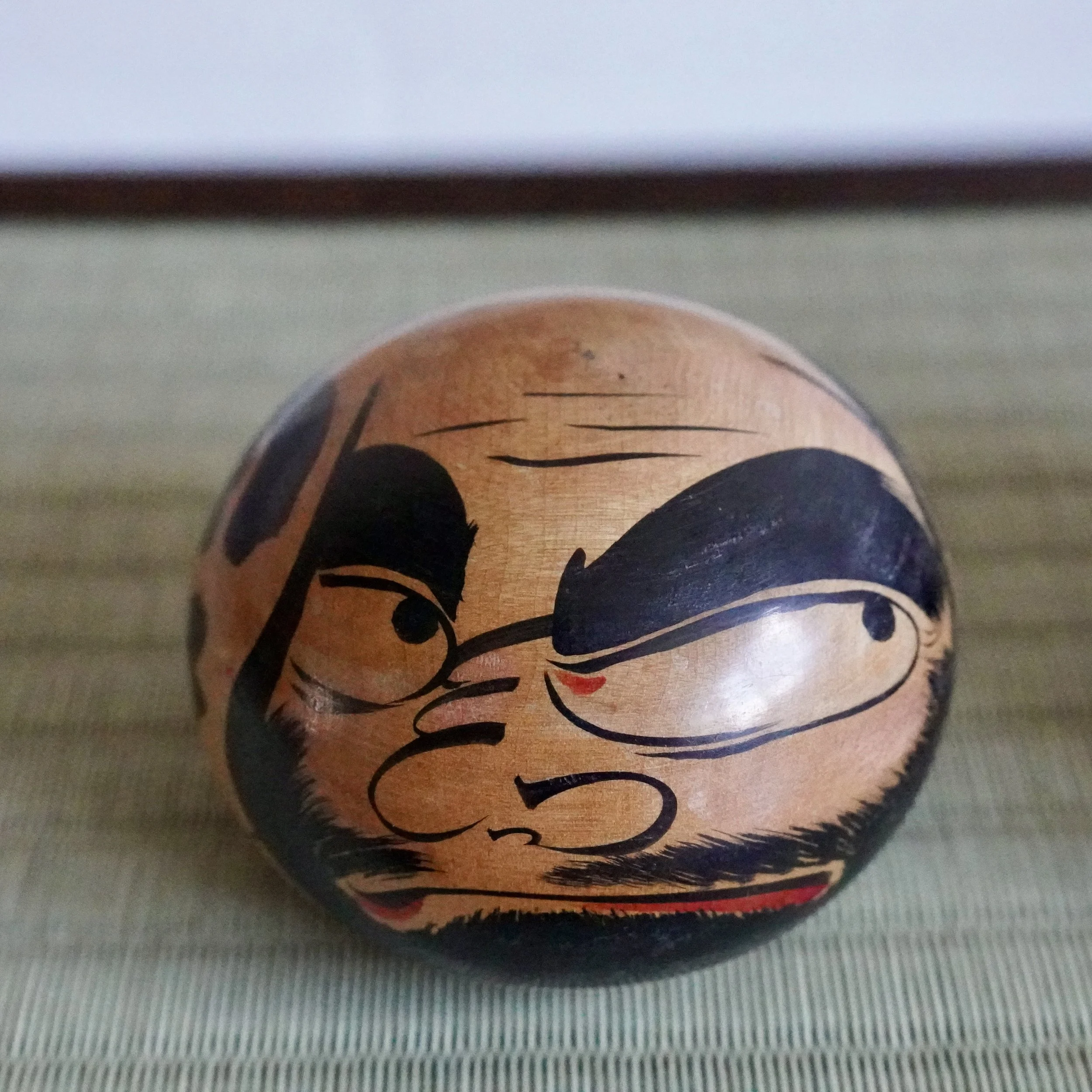 Hand-painted wooden Tsugaru daruma by Satō Zenji, showing a fierce scowling face with black brows, eyes, mustache, beard, and red mouth, with a red base band.