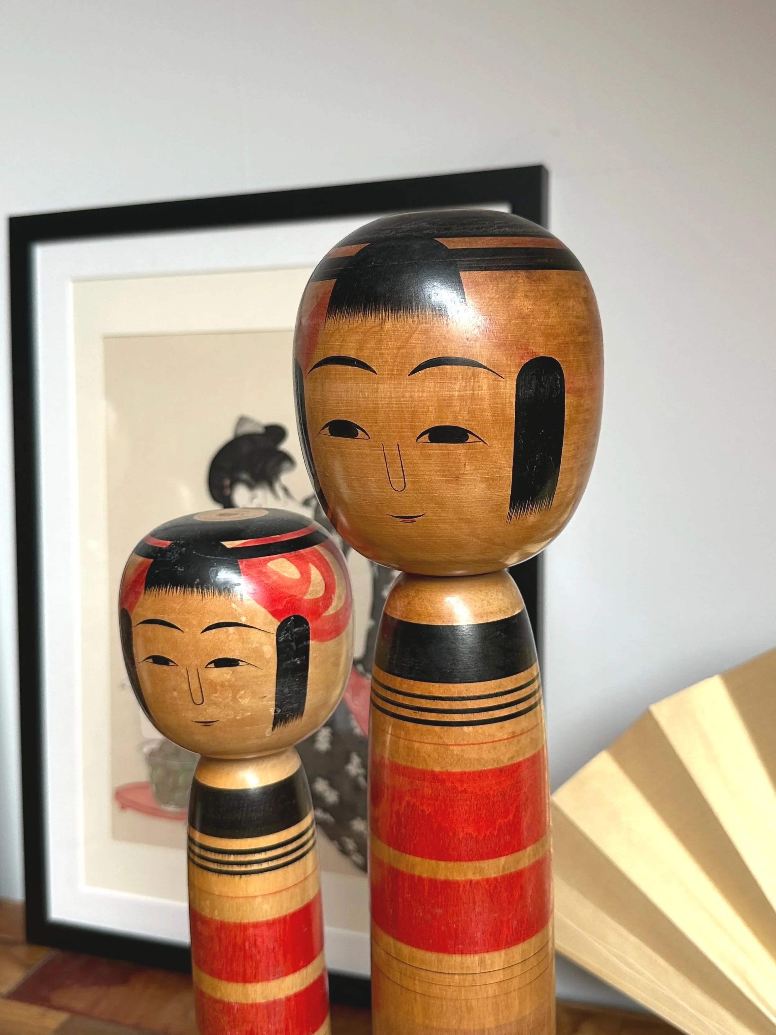 Two Tsuchiyu kokeshi dolls by Abe Ichirō, a taller doll on the right, with red, black, and natural wood banded bodies and minimalist faces.