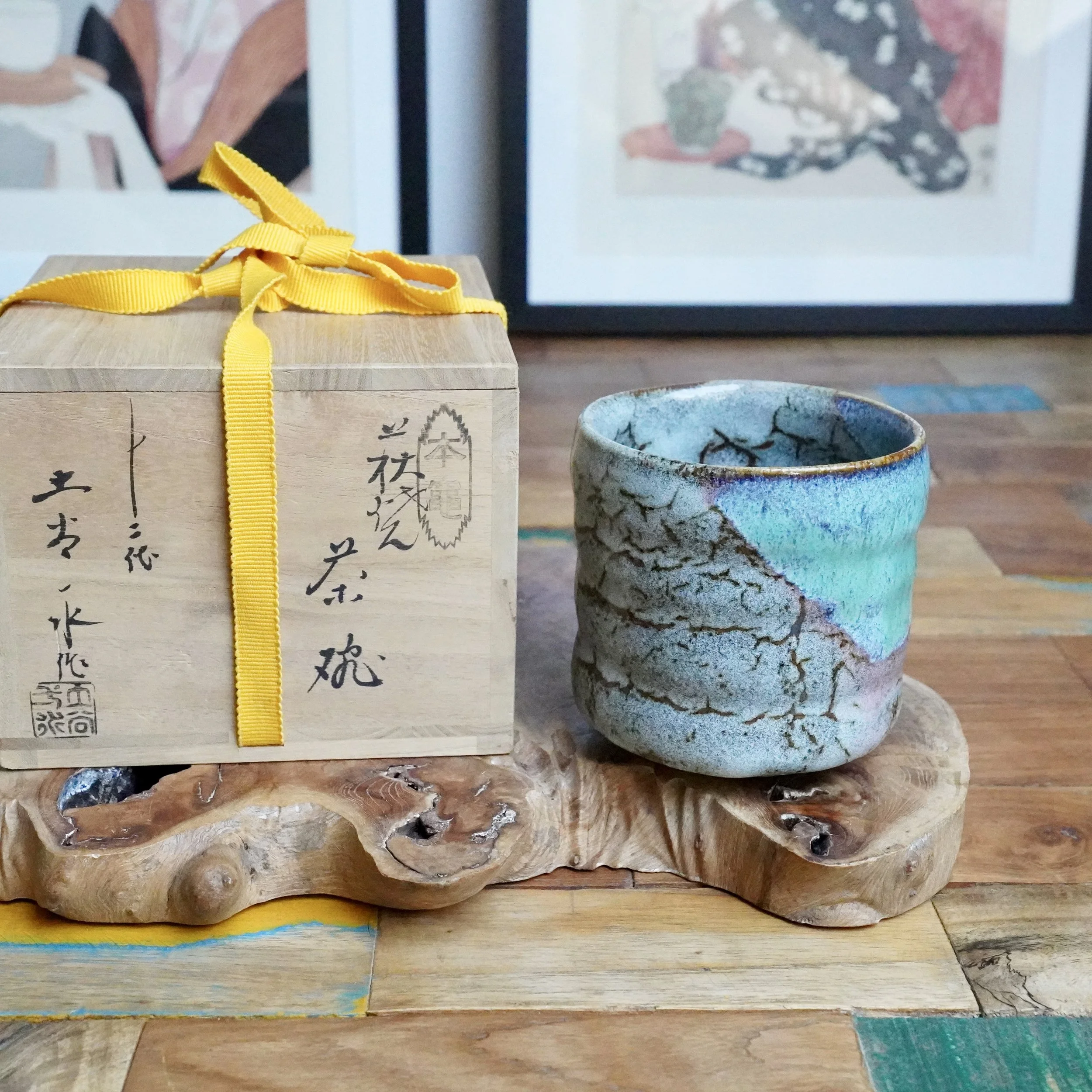 Tsutsu chawan with turquoise and grey crackle glaze, beside a wooden tomobako with Tsuchiya Issui inscription and a yellow ribbon, on a rustic wooden stand.