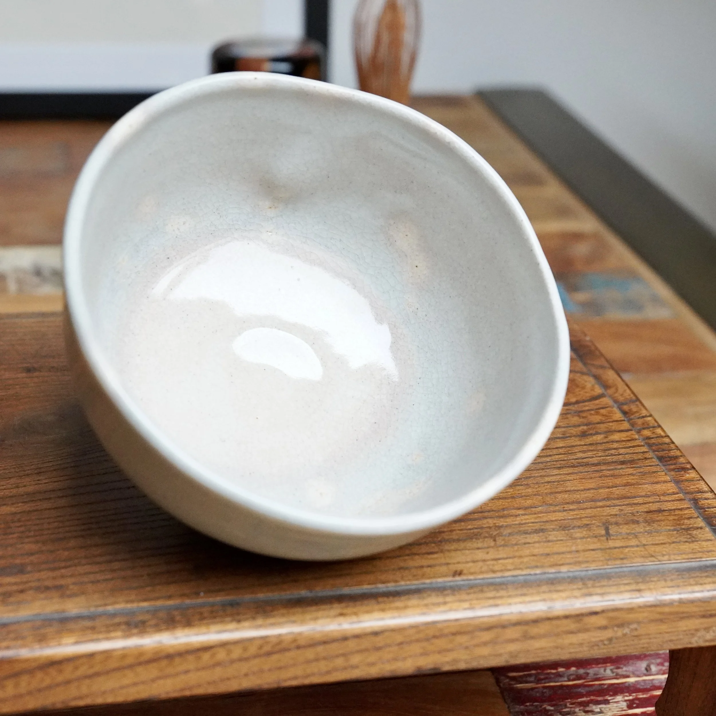 Nagasawa Minoru chawan, grey-blue crackled glaze with pink interior, subtly pressed rim, angled on a multi-toned wooden table.
