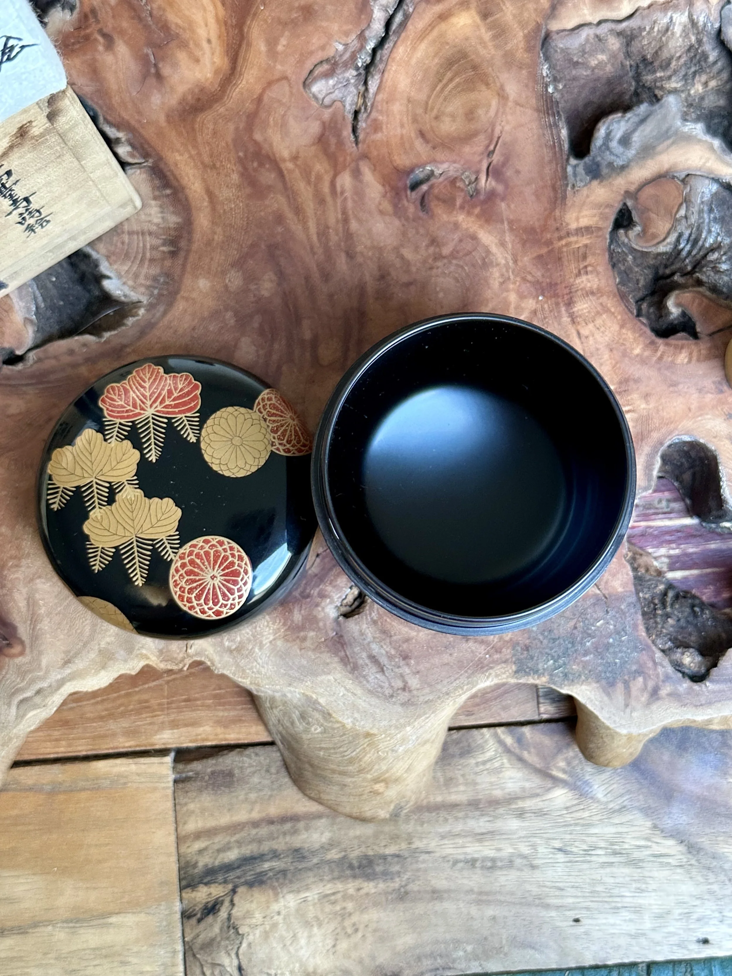 Open black lacquered natsume tea caddy and its lid with gold and red paulownia and chrysanthemum makie crests, beside a wooden box with calligraphy, on textured wood.