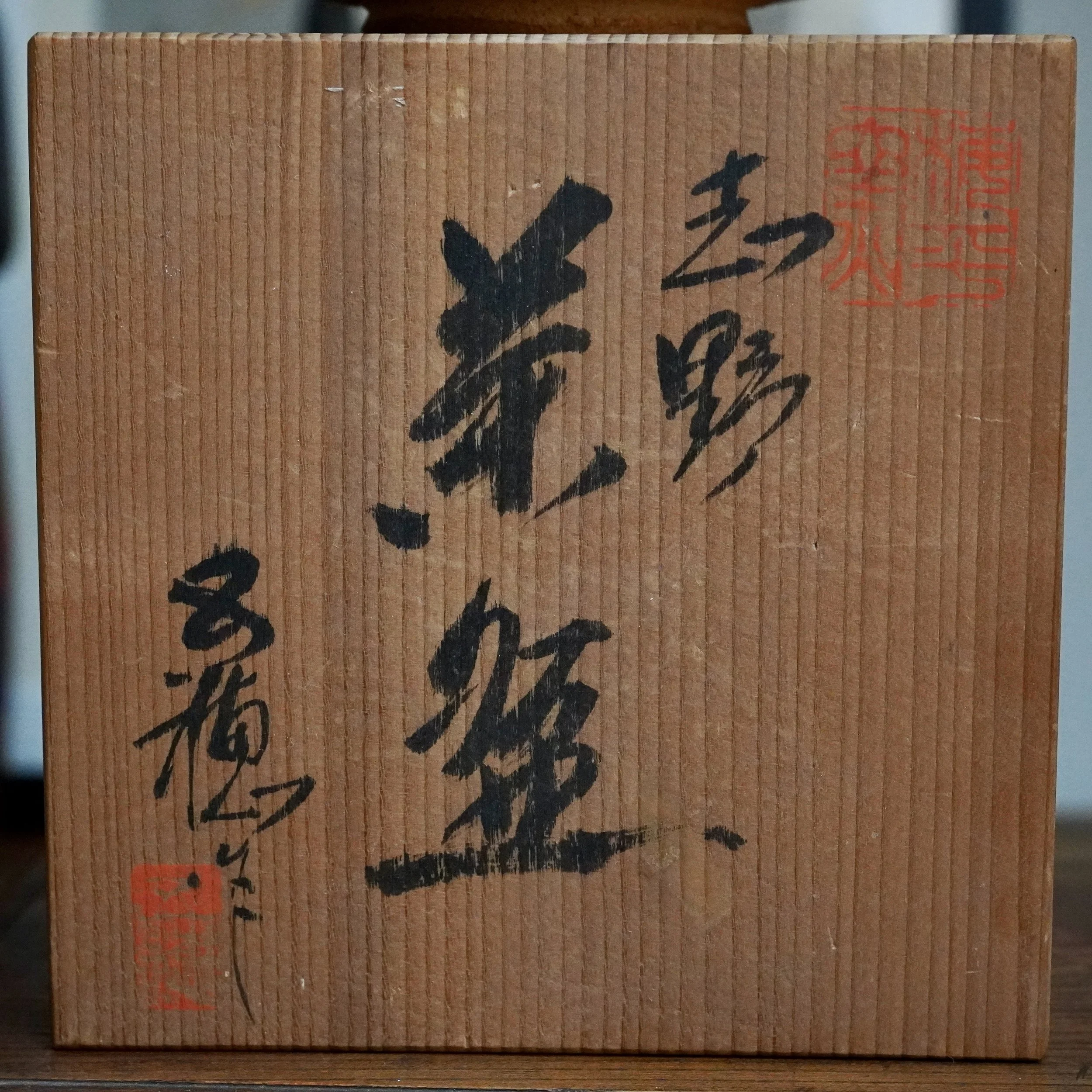 Front view of a light brown wooden tomobako with vertical grain, featuring black signed Japanese calligraphy and two red artist seals.