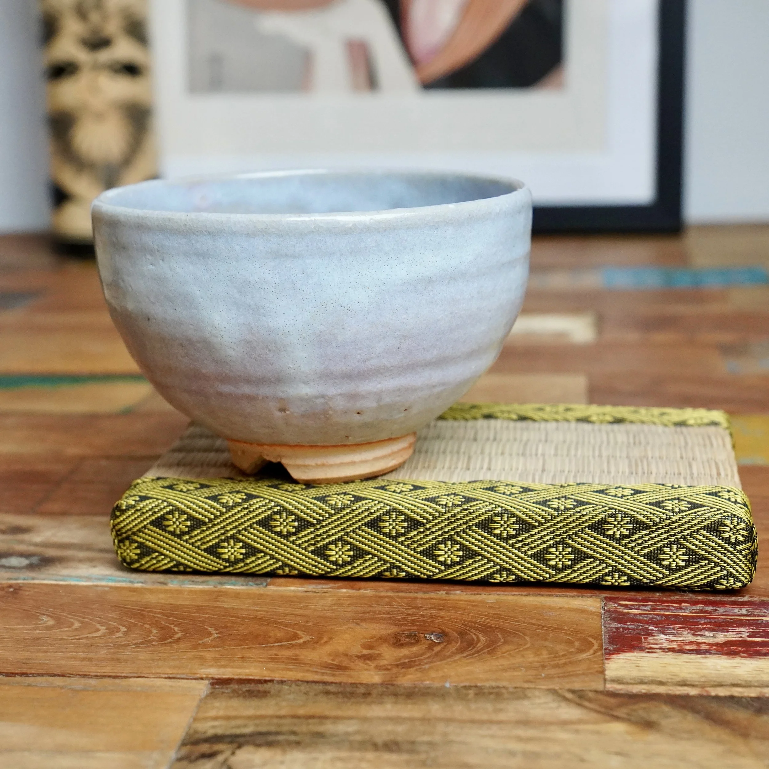 Moriwaki Fumitada's chawan, a pale speckled ceramic bowl with an unglazed reddish-brown foot, on a woven mat with yellow and black patterned trim, on a distressed wooden table.