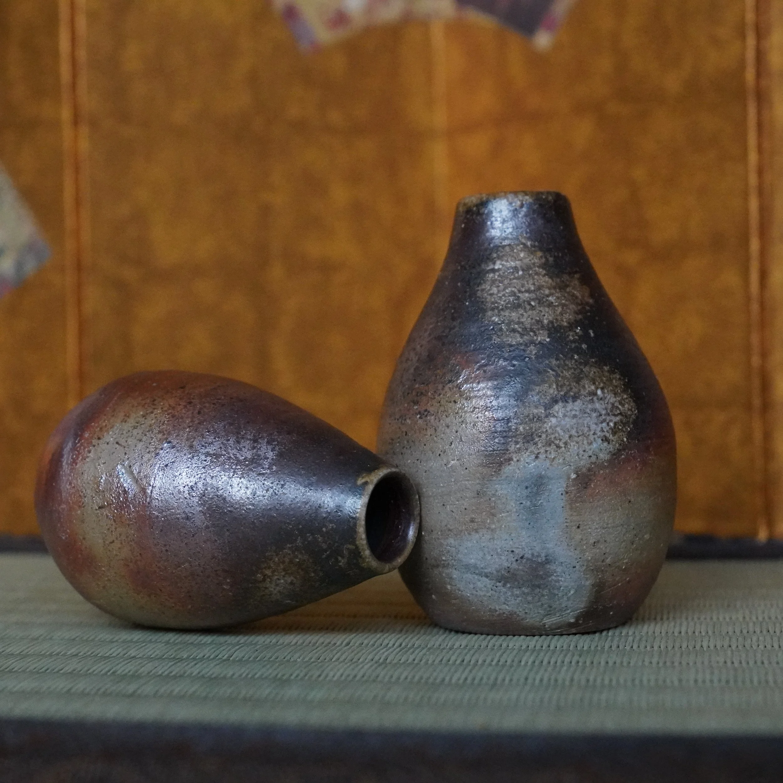 Pair of Bizen-yaki tokkuri vases by Shibaoka Tōsendō, Kōzan-gama. One upright, one tilted, showing unglazed earthy tones on a tatami mat against a gold colored screen.
