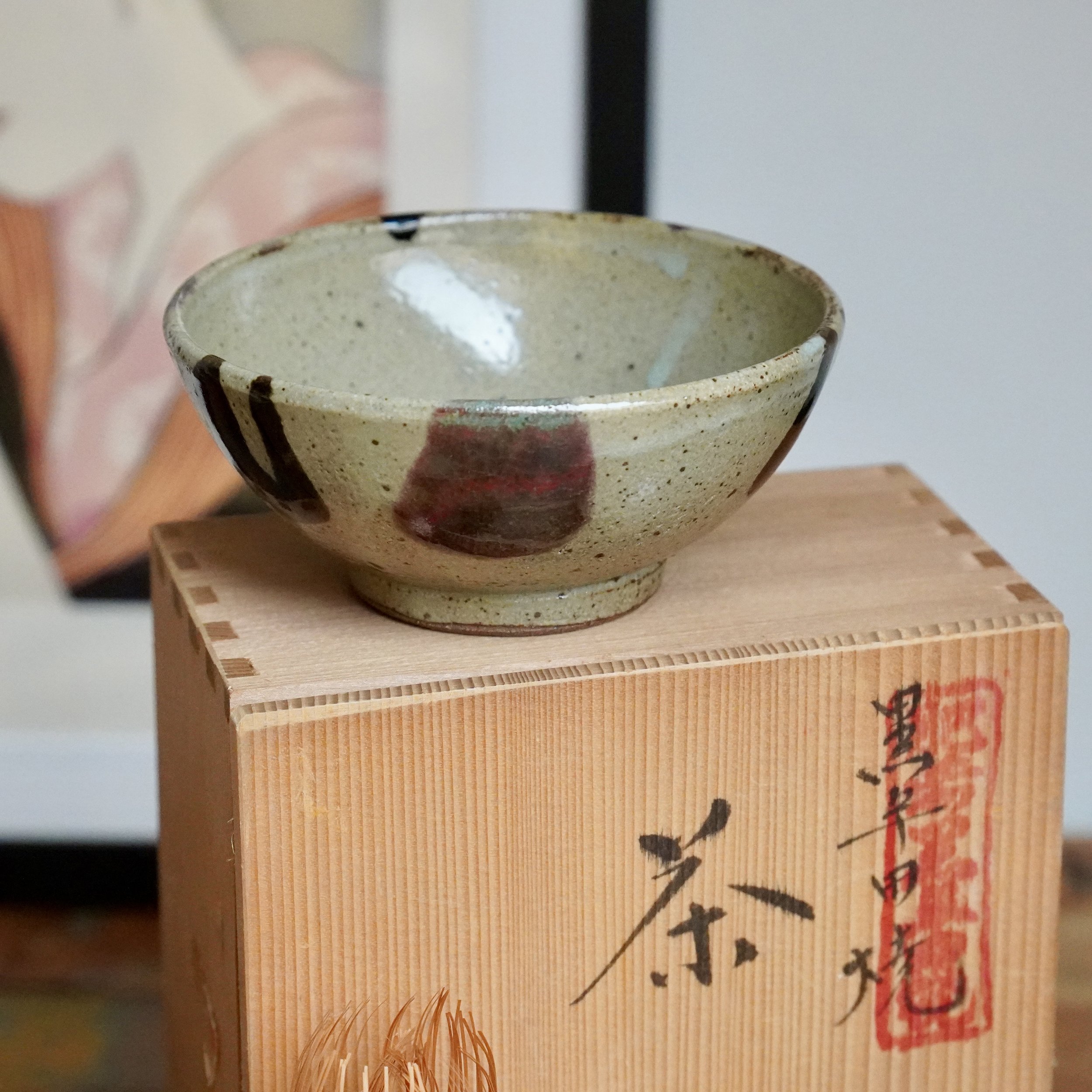 Kuromuta-yaki hira-chawan, by Maruta Nobumasa kiln, with speckled pale glaze, dark strokes, and a red patch, on a wooden tomobako with calligraphy and a red seal.