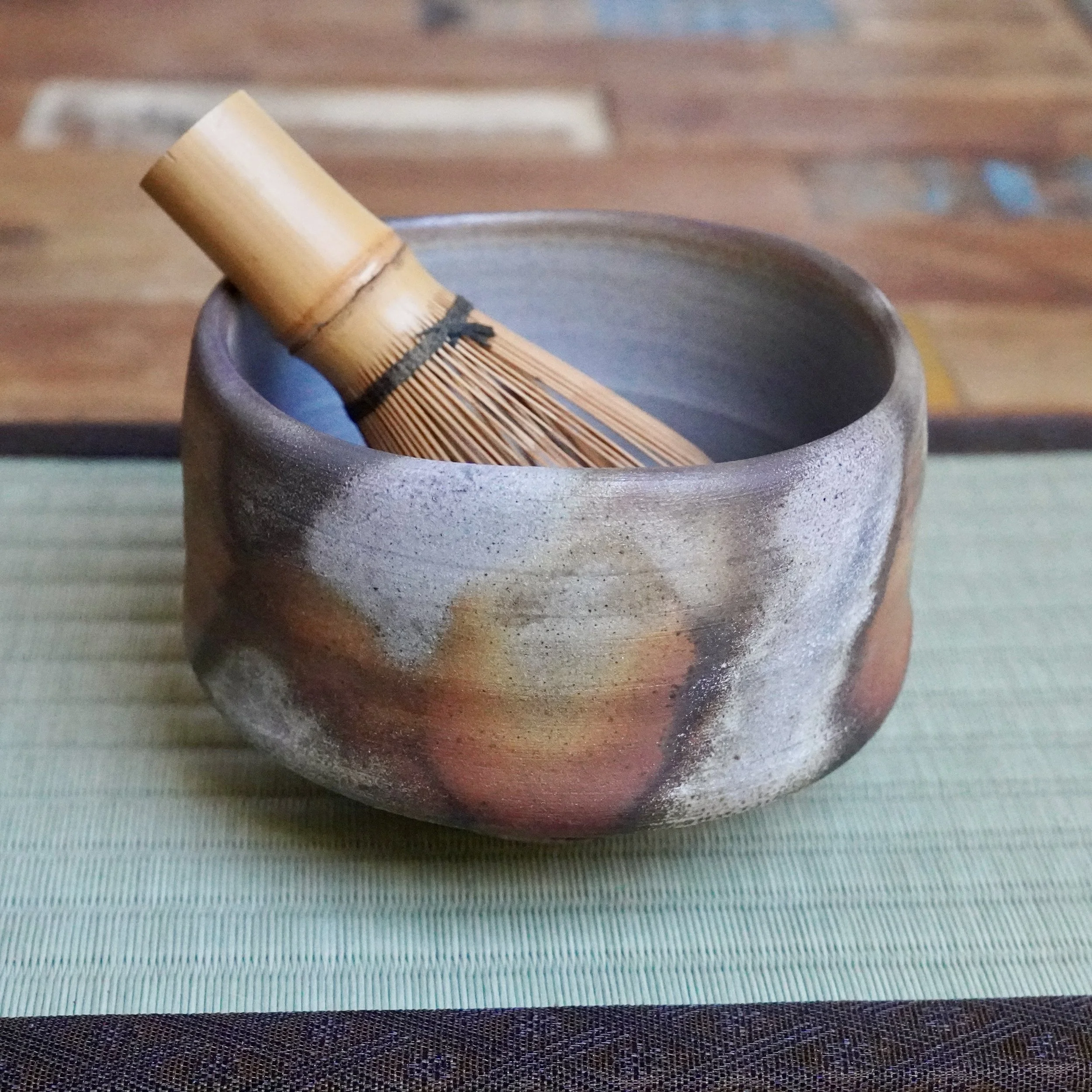 Inomata Masaaki Sangiri Bizen chawan with rustic grey, rust, and orange firing patterns, holding a bamboo matcha whisk. The unglazed bowl sits on a tatami mat.
