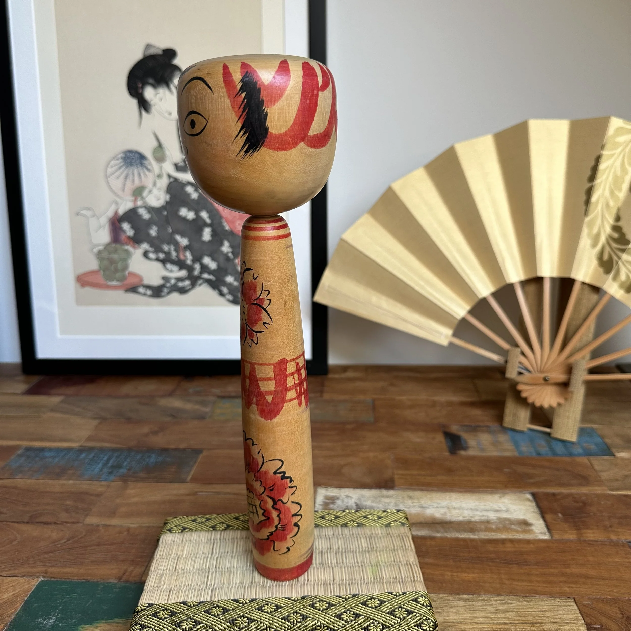 Seya Jūji's Nakanosawa Takobozu kokeshi doll, with a large head featuring painted black eyes and bold red patterns, atop a slender body adorned with red cherry blossoms, on a woven mat.