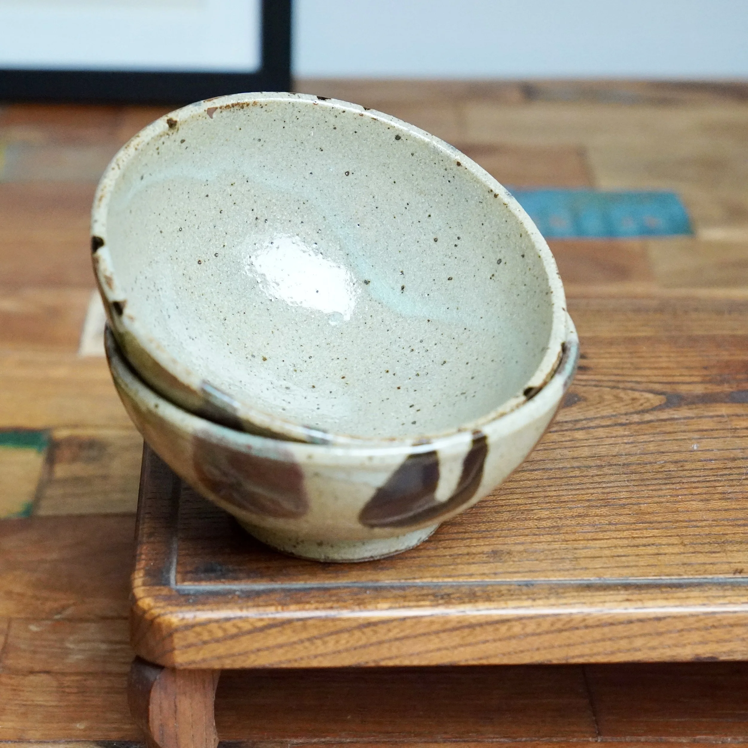 Two speckled, light-colored hira-chawan bowls stacked on wood. The top bowl is angled, revealing its glossy interior and dark, irregular glaze marks.