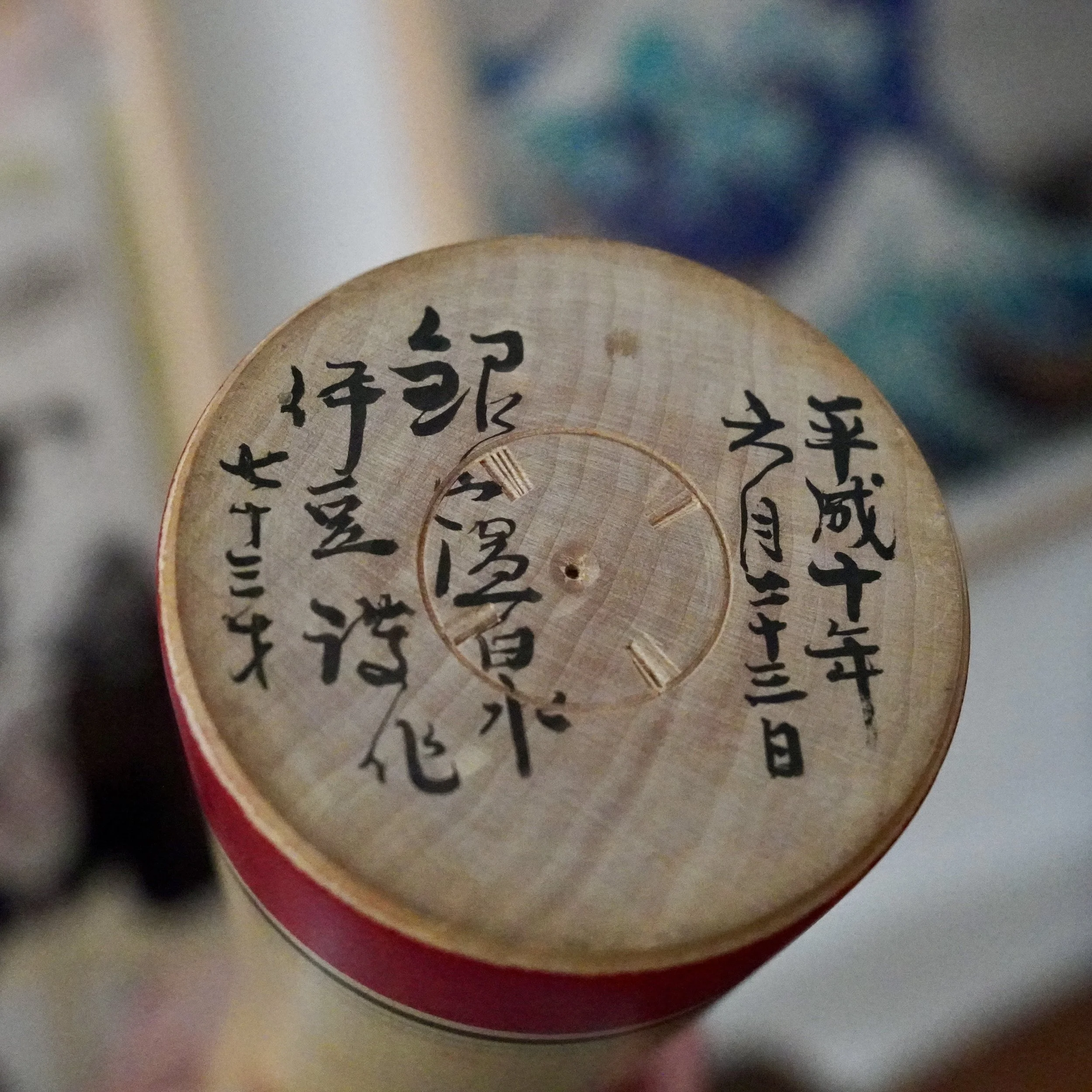 Underside of a Ginzan kokeshi by Izu Mamoru, with black calligraphy on a light wood base with a red rim, dated June 23, 1998, and age 73.