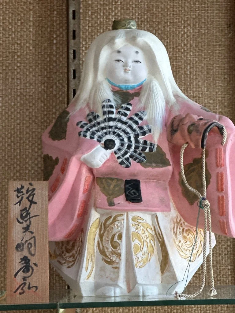 A traditional Japanese ceramic statue of a vintage joyful, kabuki figure with a round face, long white hair, dressed in ornate pink and gold robes, holding a fan, with a wooden plaque with Japanese calligraphy in front.