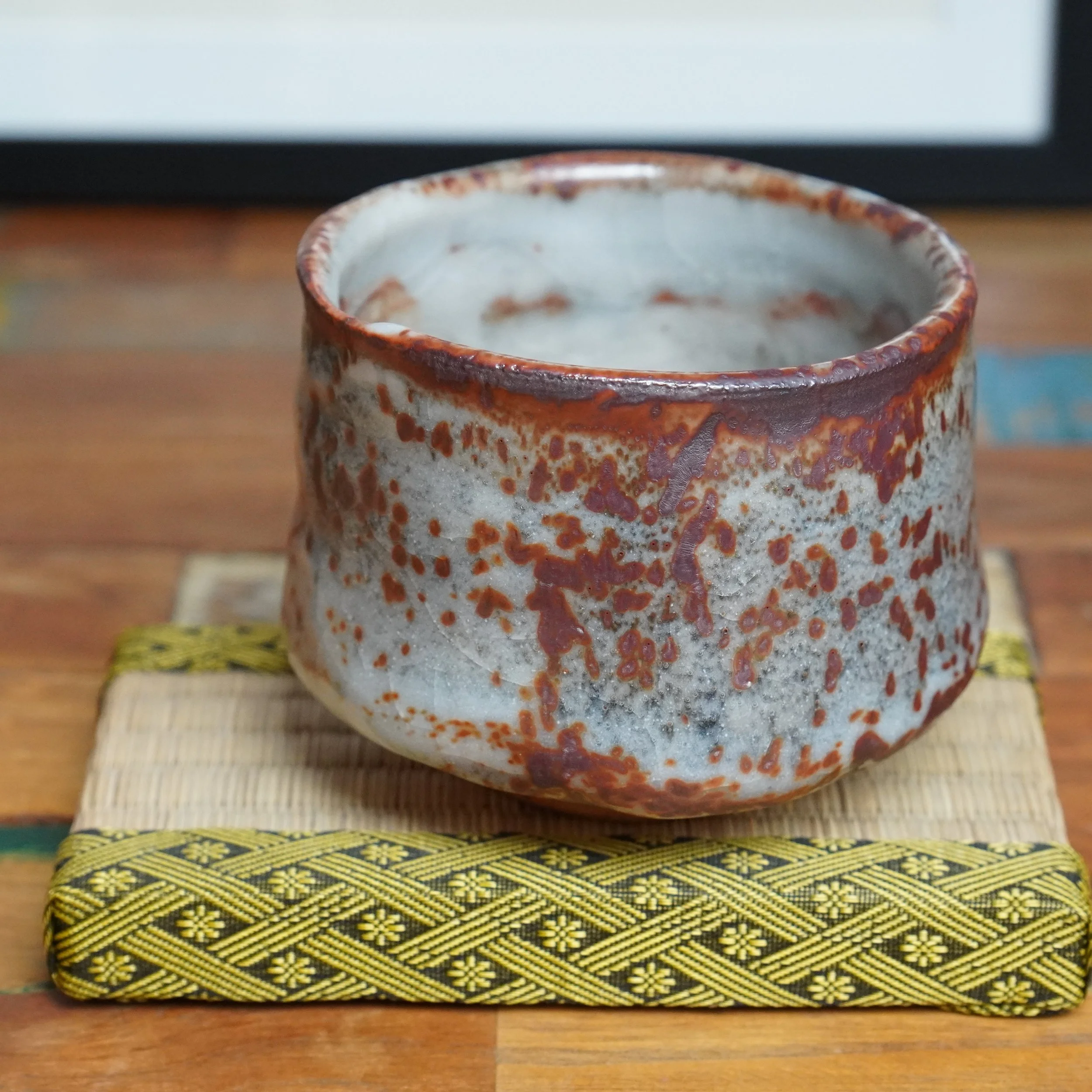 Shino chawan, pale grey with irregular reddish-brown speckles and drips, rests on a woven mat with a yellow and black geometric border.