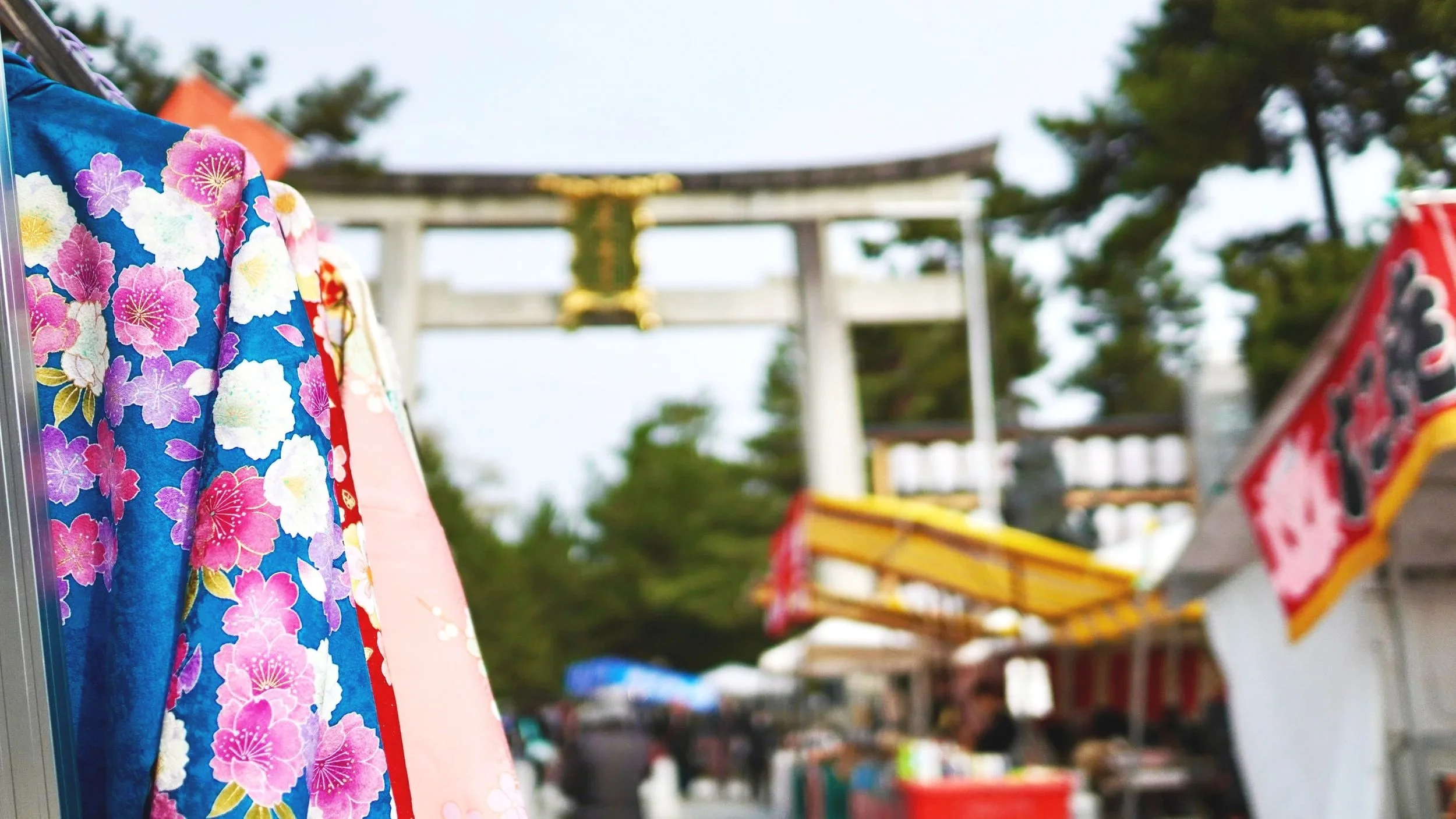 A vibrant scene at the Tenjin-san antique market at Kitano Tenmangu Shrine in Kyoto, featuring colorful vintage silk kimonos with floral patterns hanging in the foreground, with the large stone torii gate and market stalls blurred in the background.