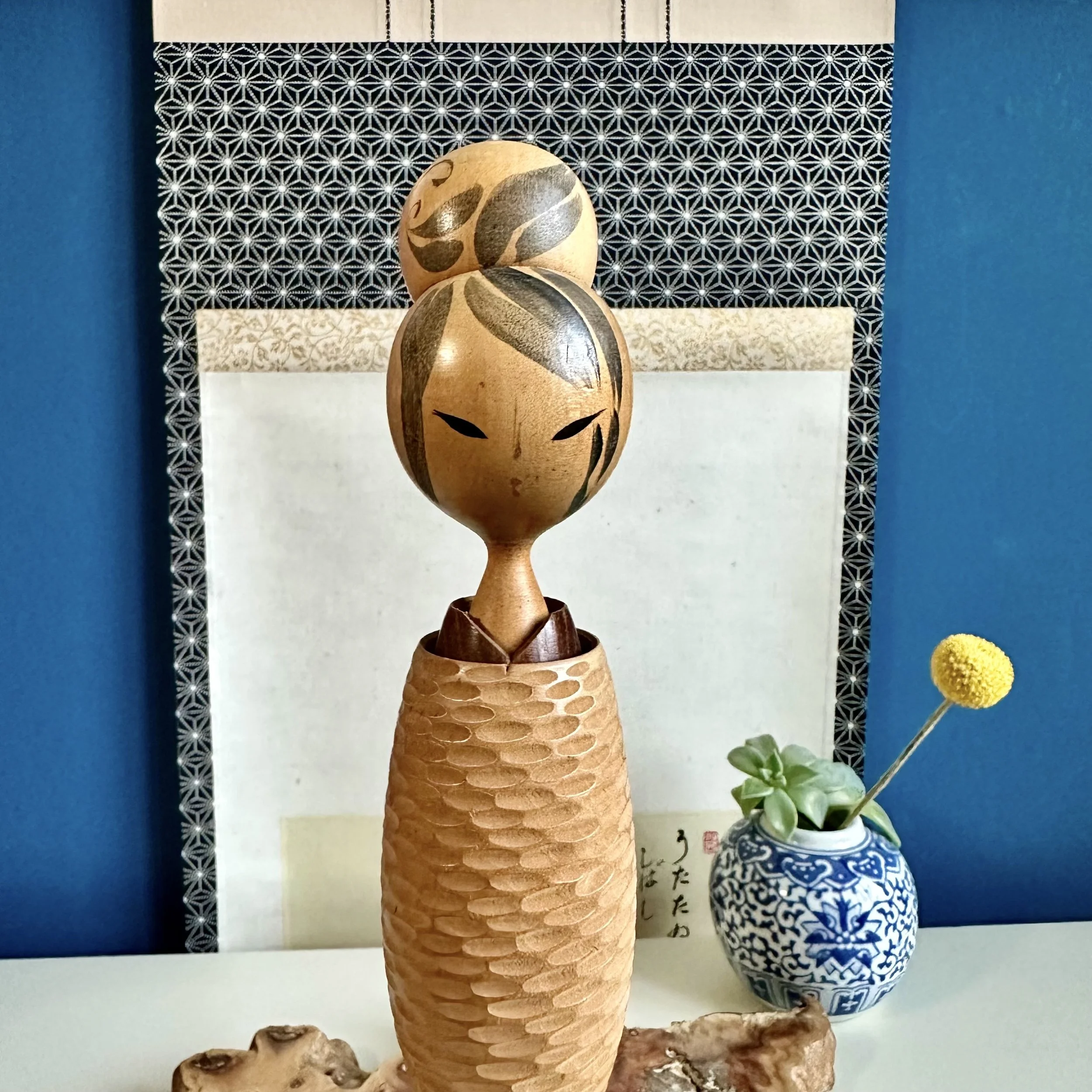 A carved wooden kokeshi creative with a Japanese calligraphy scroll and patterned fabric in the background, along with a potted plant with yellow flowers.