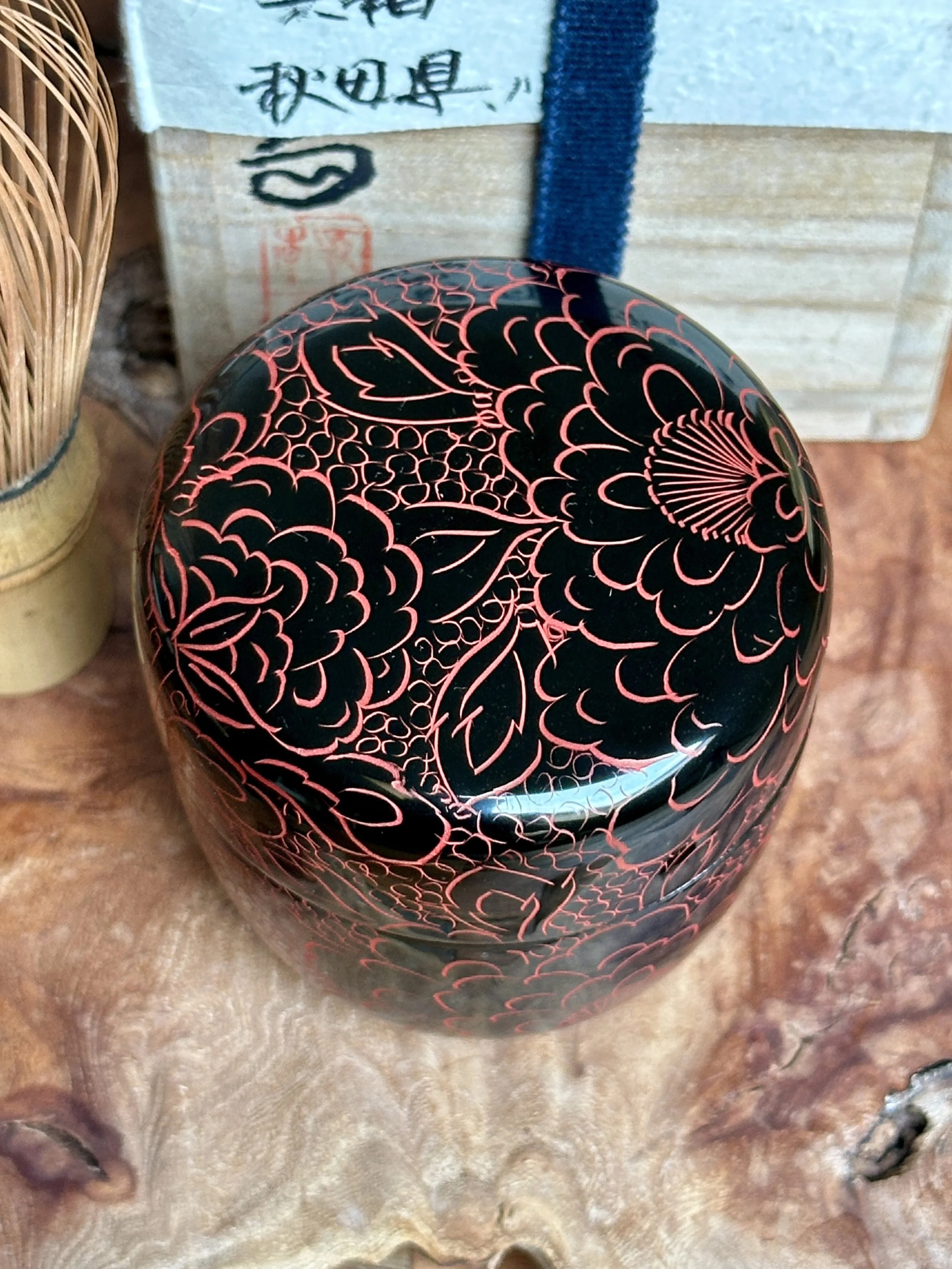 Rounded black lacquer tea caddy, signed Fujiwara Seiu, featuring glossy vermilion engraved peony and delicate floral patterns, seen from above.
