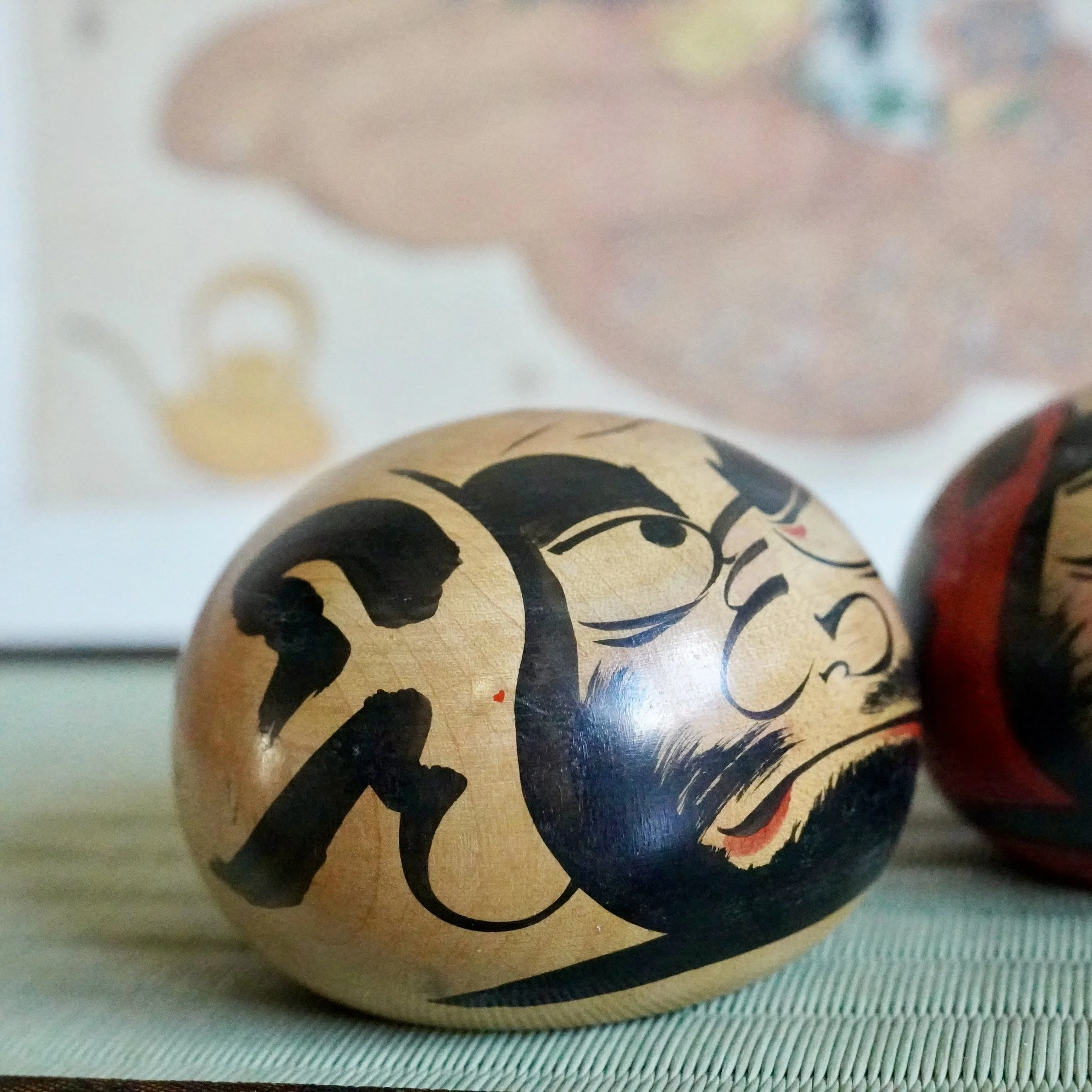 Wooden Tsugaru daruma by Satō Zenji with fierce black painted features, calligraphic text, and a small red mouth accent, on a textured mat. Another daruma is blurred in the background.