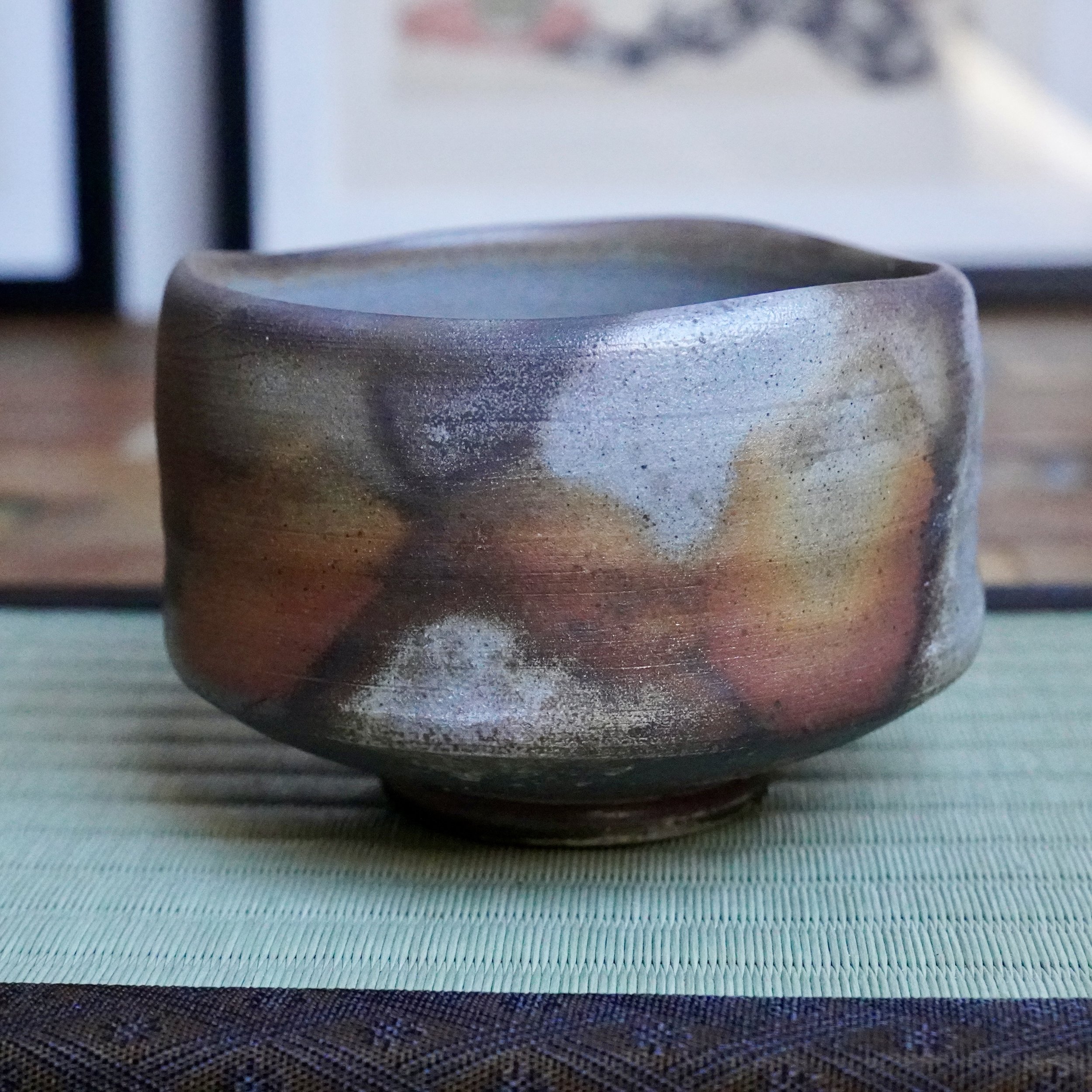 Inomata Masaaki's Sangiri Bizen chawan, unglazed with wood-fired marks in dark brown, rust orange, and ash grey, with a silvery rim, on a green mat.