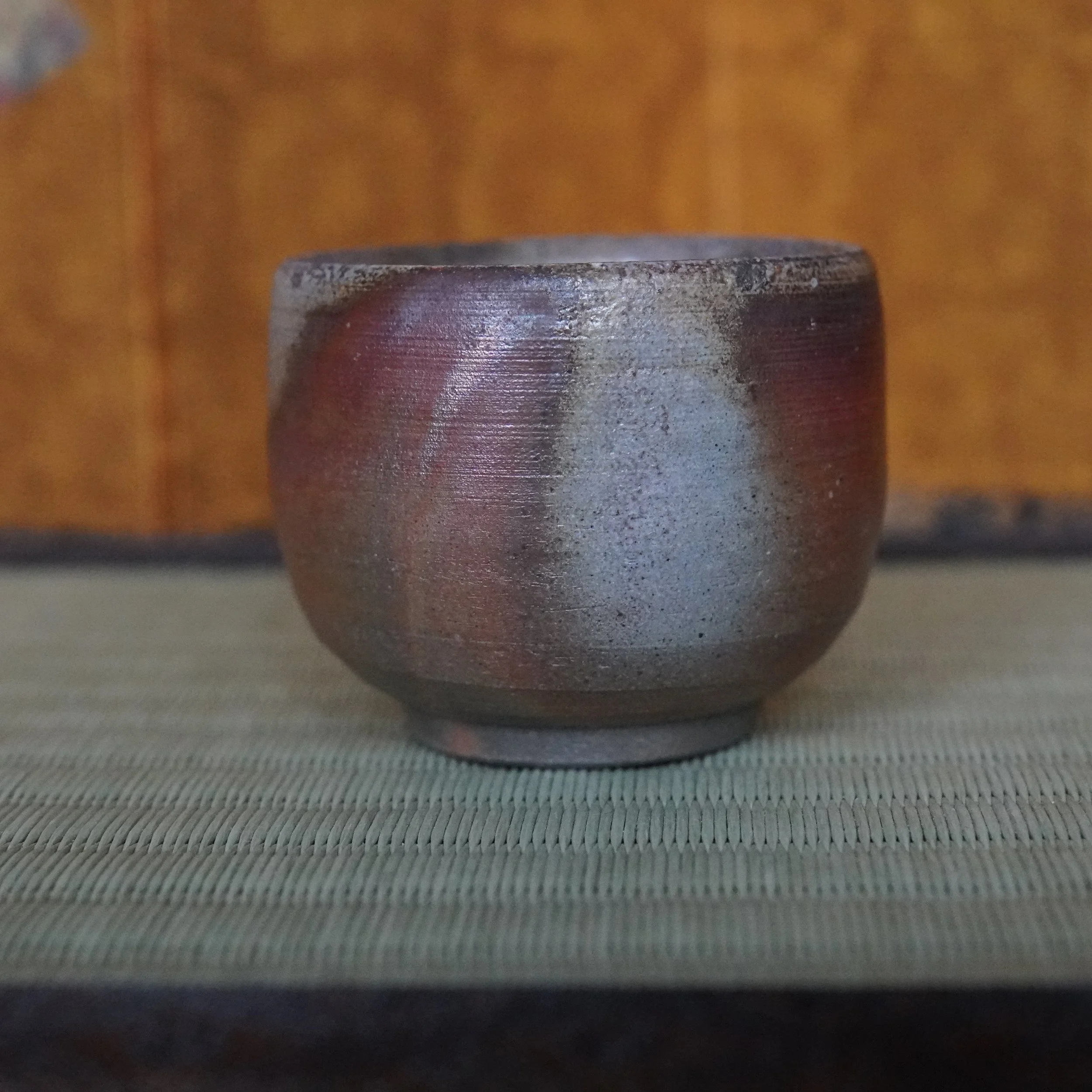 Bizen-yaki guinomi with distinct scarlet hidasuki flame marks over mottled gray and ochre, on a woven tatami mat.
