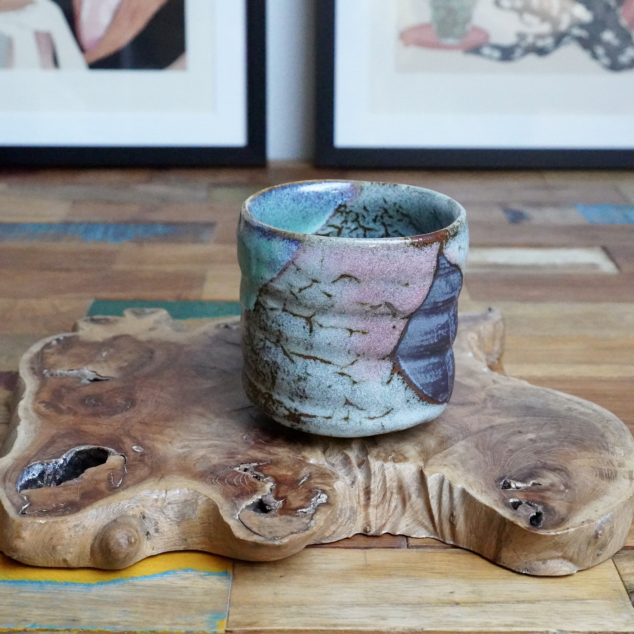 Hand-thrown tsutsu chawan with turquoise, grey, and lilac glaze and warm brown crackle lines, on an organic wood slab.