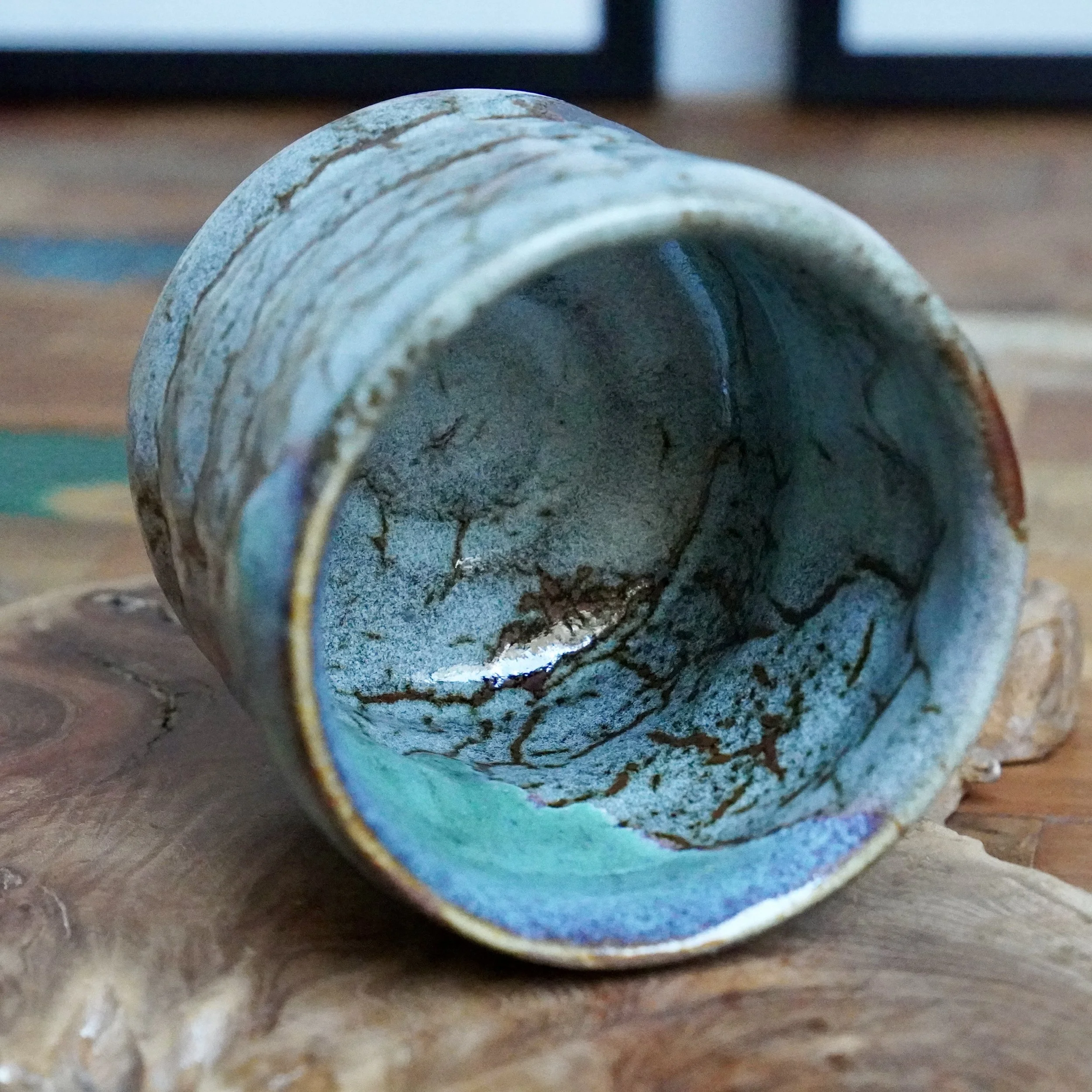 Hand thrown tsutsu chawan, angled to reveal its interior. Features layered turquoise and speckled grey glaze with warm brown lines and a vibrant blue rim, resting on wood.