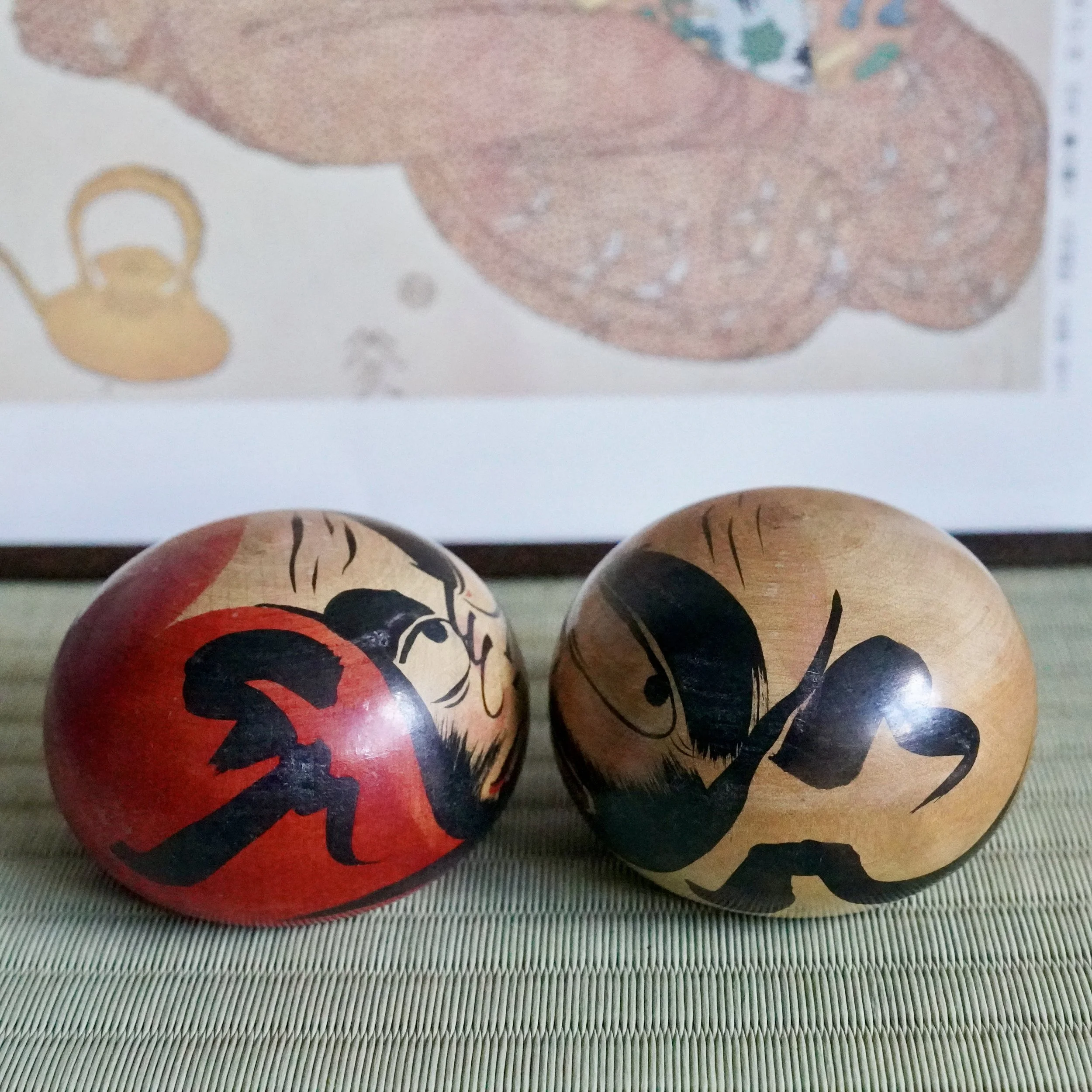 Satō Zenji, Nuruyu Tsugaru daruma pair. One red and black, one natural wood and black, hand-painted scowling faces on a tatami mat.
