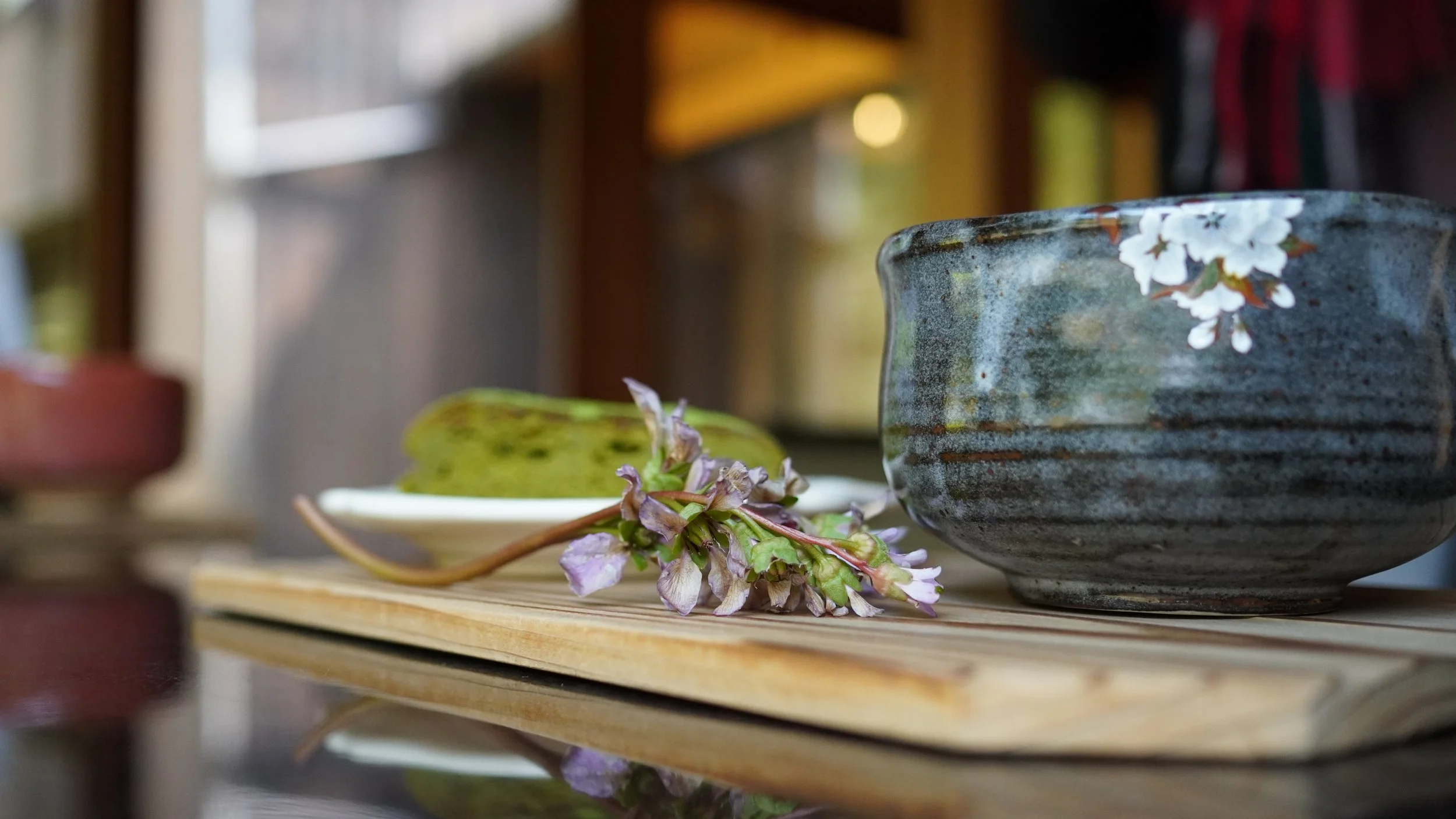 Dark-glazed vintage Japanese chawan on a wooden tray with cherry blossom branches and a green wagashi sweet. Matcha bowl for the tea ceremony, personally sourced in Japan.