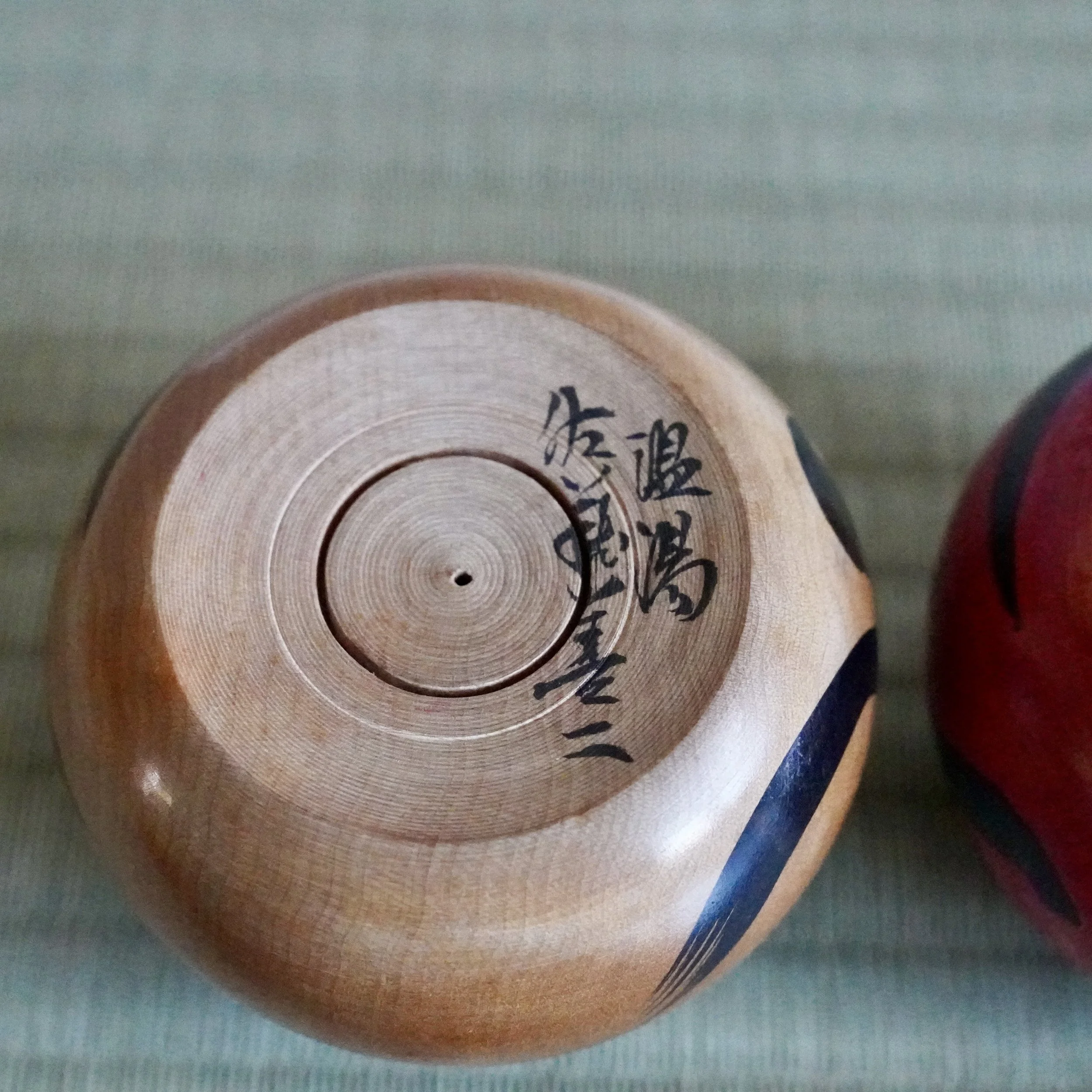 Underside of a wooden daruma, revealing lathe marks and black calligraphy for Nuruyu and Satō Zenji. A black stripe is on its side, with a red daruma partly visible on the right.