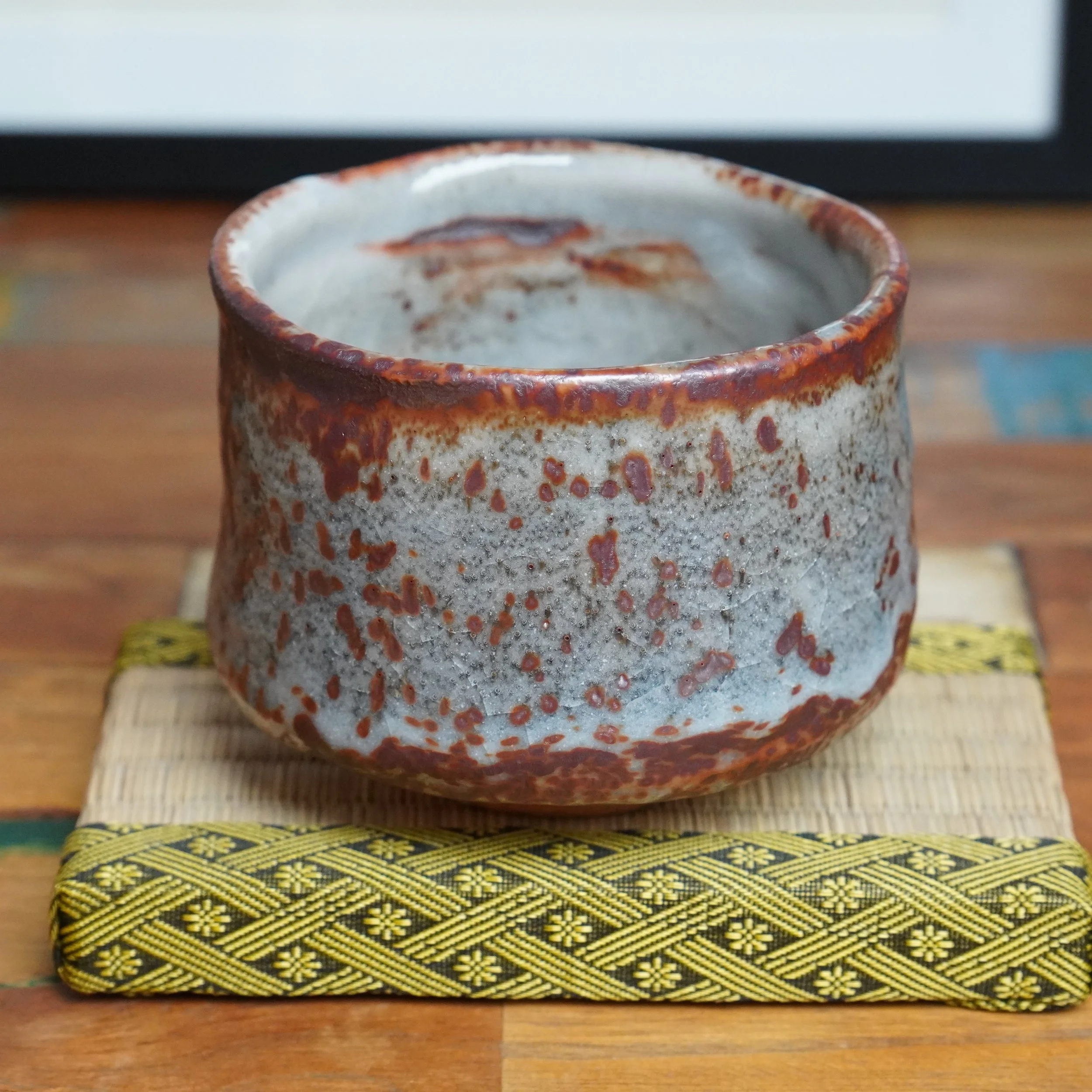 Ceramic chawan with a light grey, crazed glaze base heavily dappled with deep red speckles and blotches, topped by a reddish-brown rim.