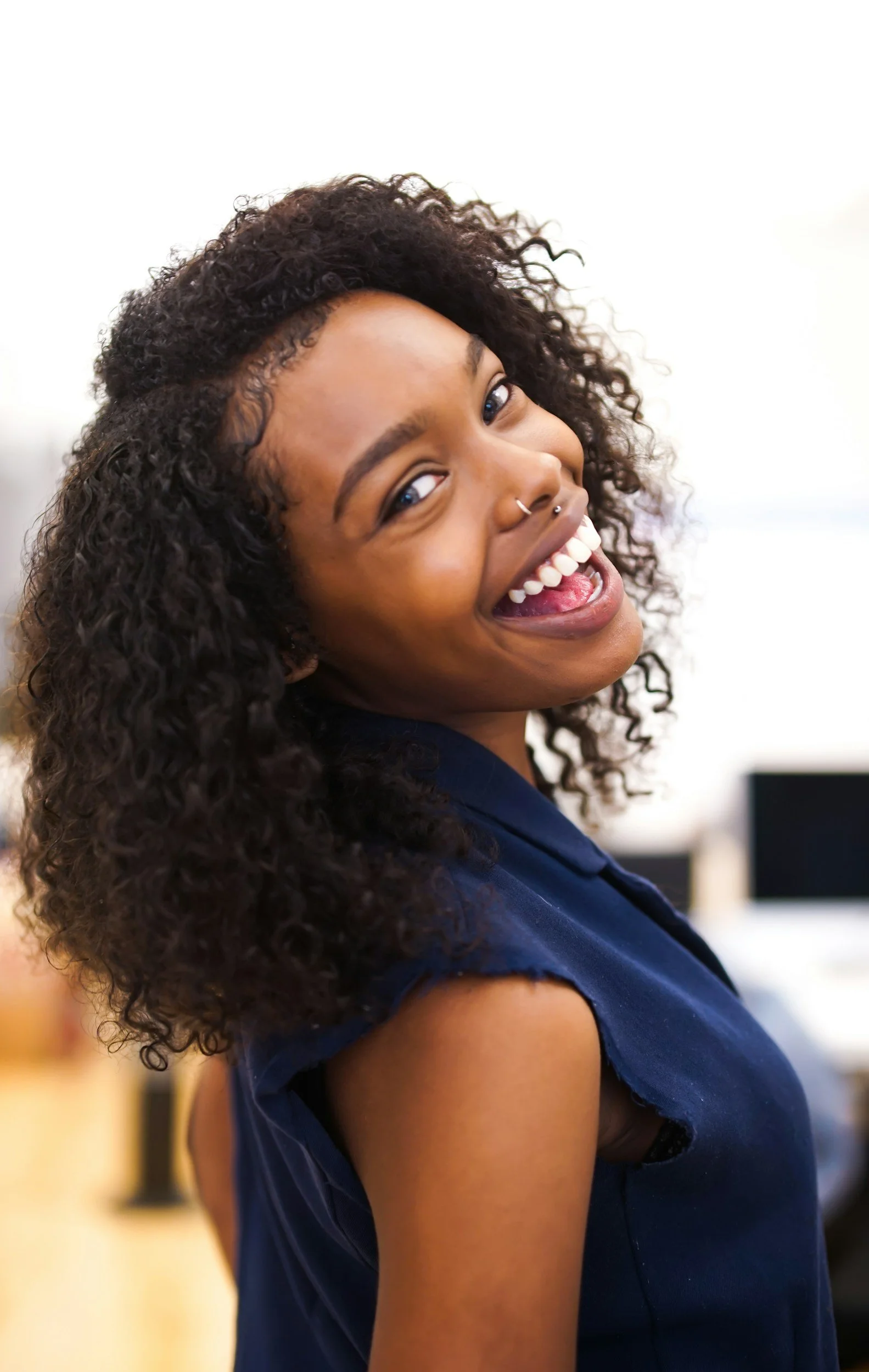 A young woman with curly hair, nose piercings, and a wide smile, wearing a sleeveless navy blue top, looking back at the camera in an indoor setting.
