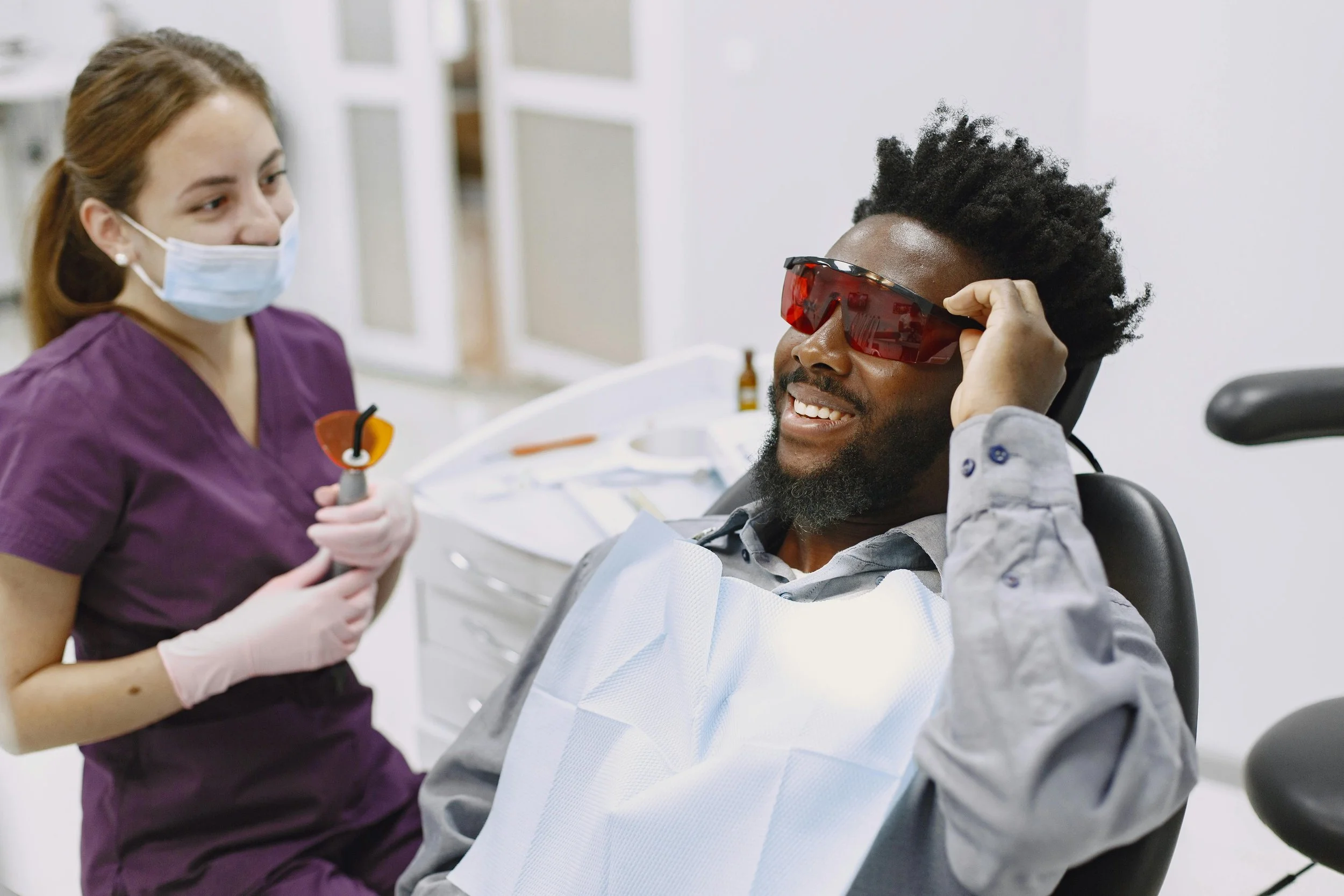 A man with a beard and glasses is sitting in a dental chair, smiling, wearing protective red glasses and a bib. A female dental professional in scrubs and a face mask is holding dental tools while talking to him.
