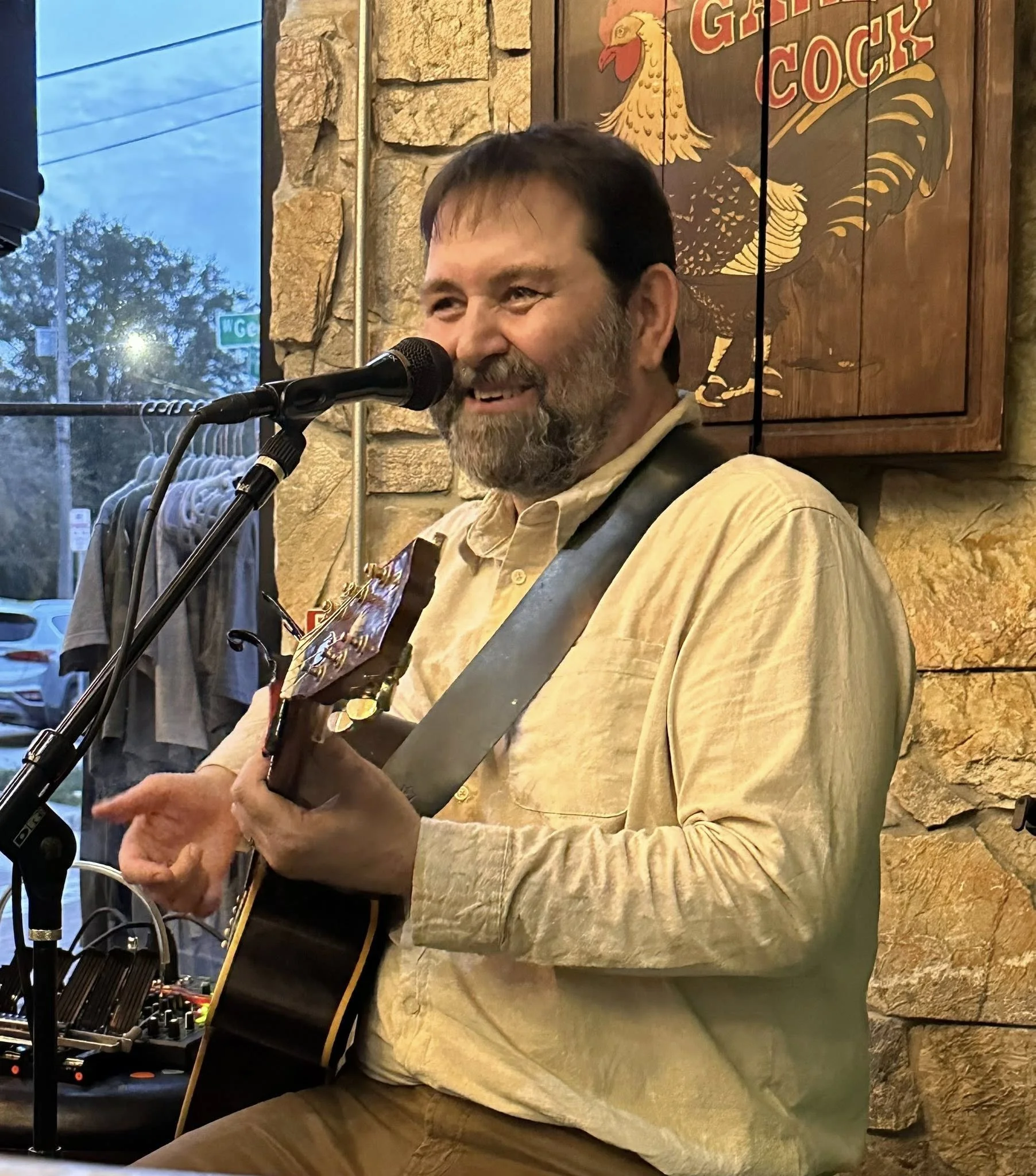 A man with a beard and mustache singing into a microphone while playing an acoustic guitar in a cozy venue with stone walls and a sign behind him that reads 'Giant Cock'.
