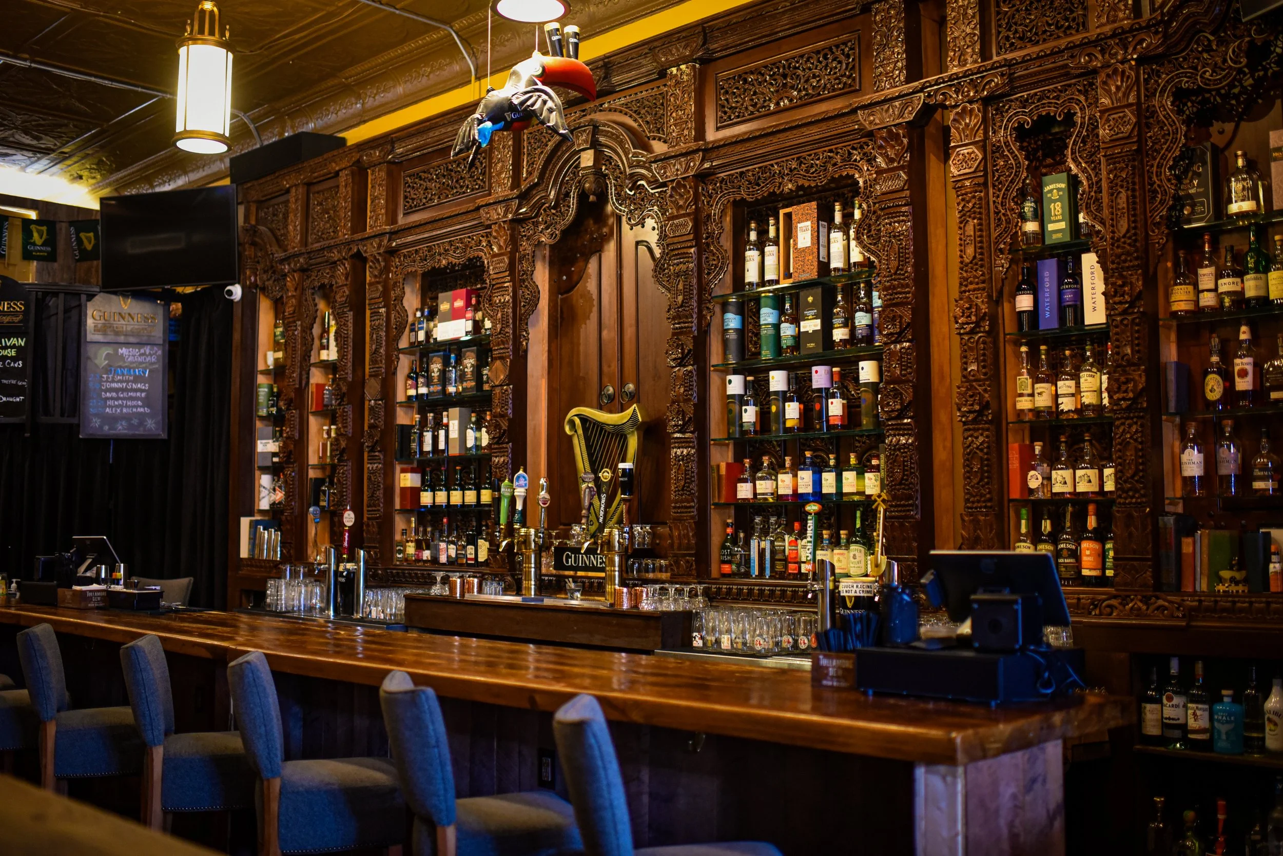 Inside a bar with dark wood and ornate carved wood panels, a wooden bar counter with grey upholstered chairs, and shelves filled with various bottles of liquor. There is a horse head mounted on the wall, beer taps on the counter, a TV mounted on the wall, and a chalkboard sign.