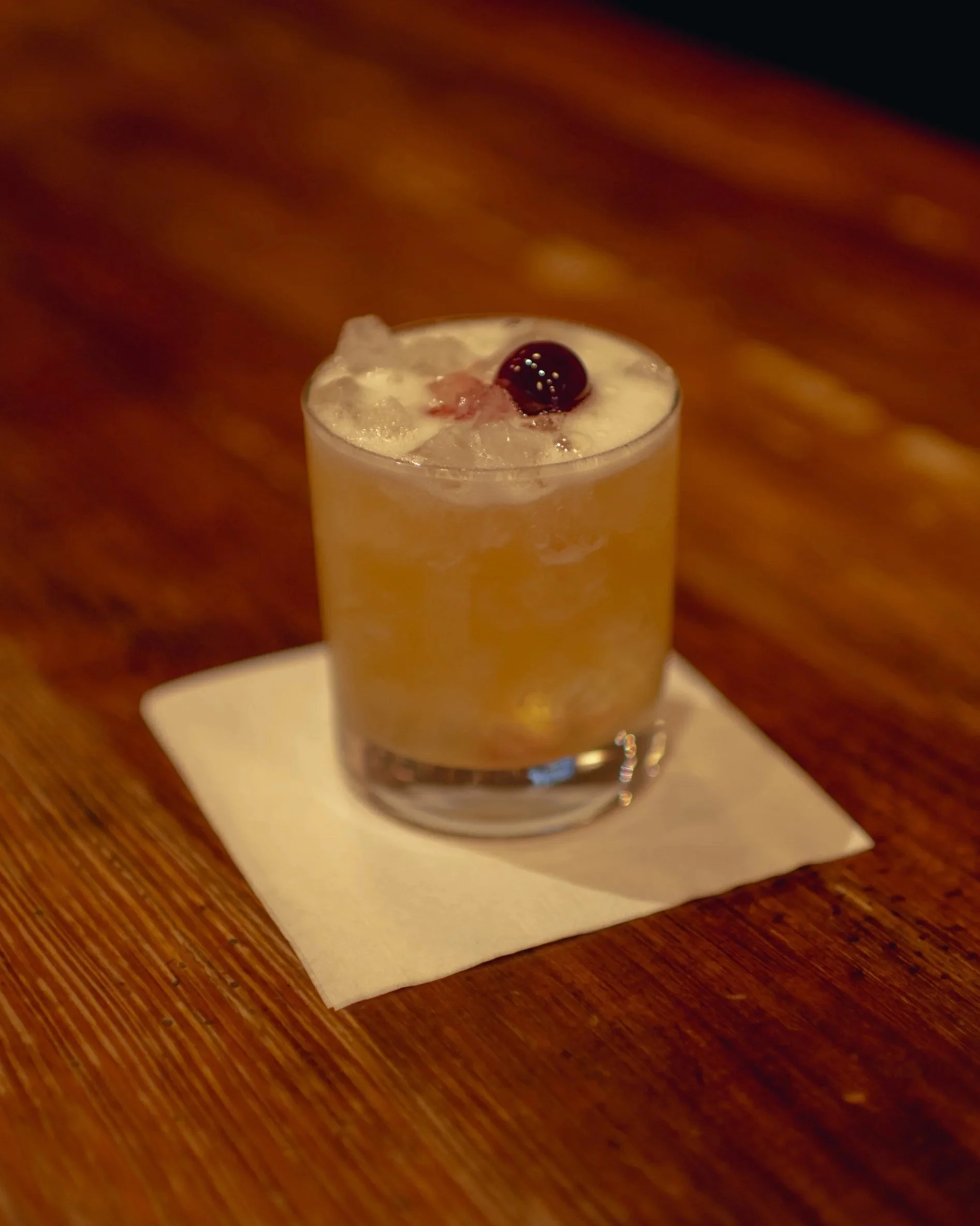 A cocktail drink with ice and a cherry on top on a white napkin on a wooden table.