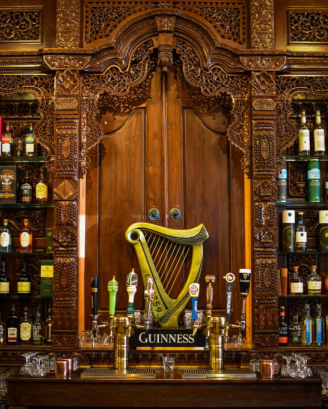 Intricately carved dark wood bar with Guinness harp logo, several beer taps, liquor bottles on shelf, and glasses underneath.