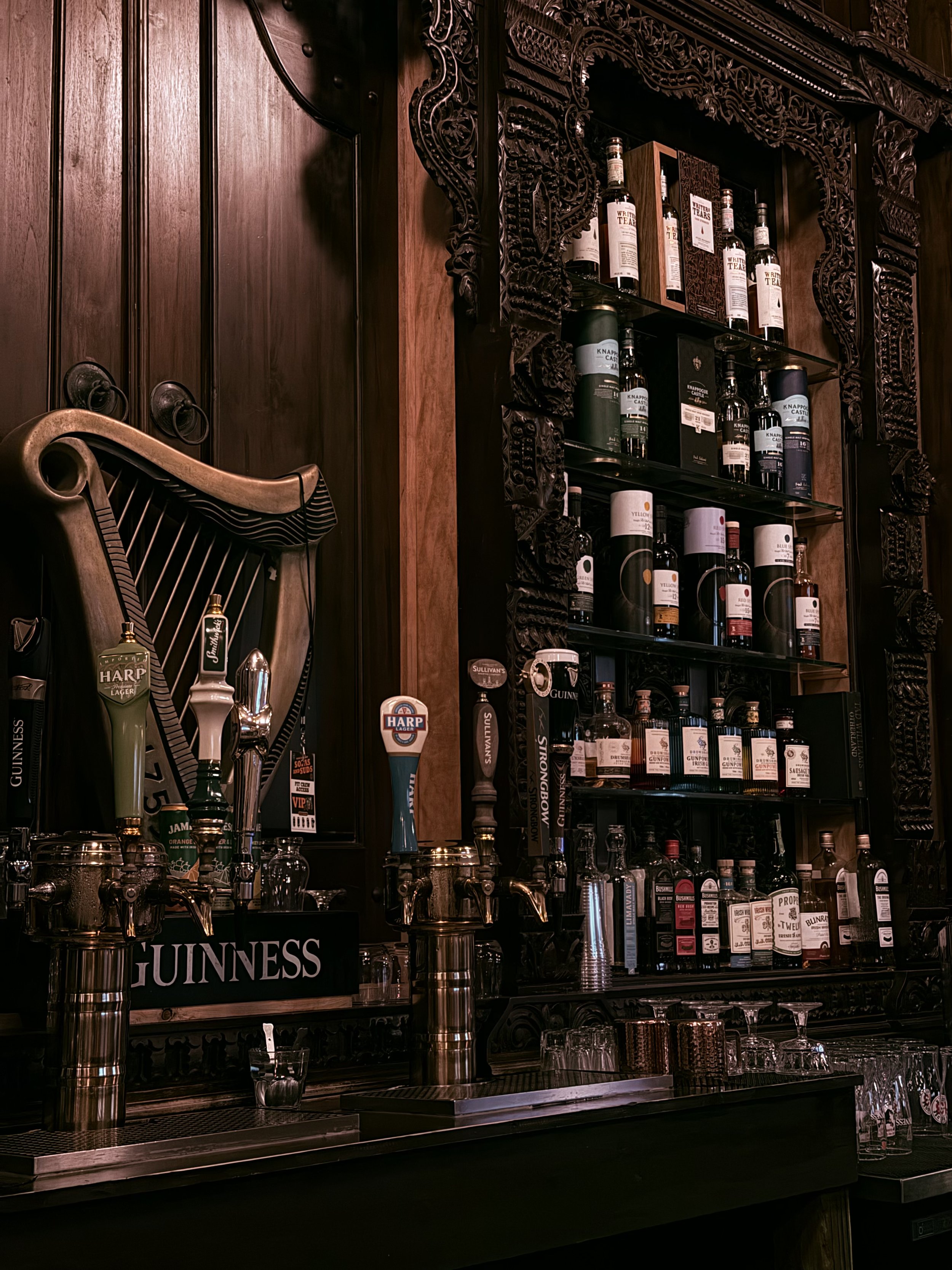 View of a bar with wooden decor, multiple bottles of liquor on shelves, and beer taps labeled Guinness and Harp.