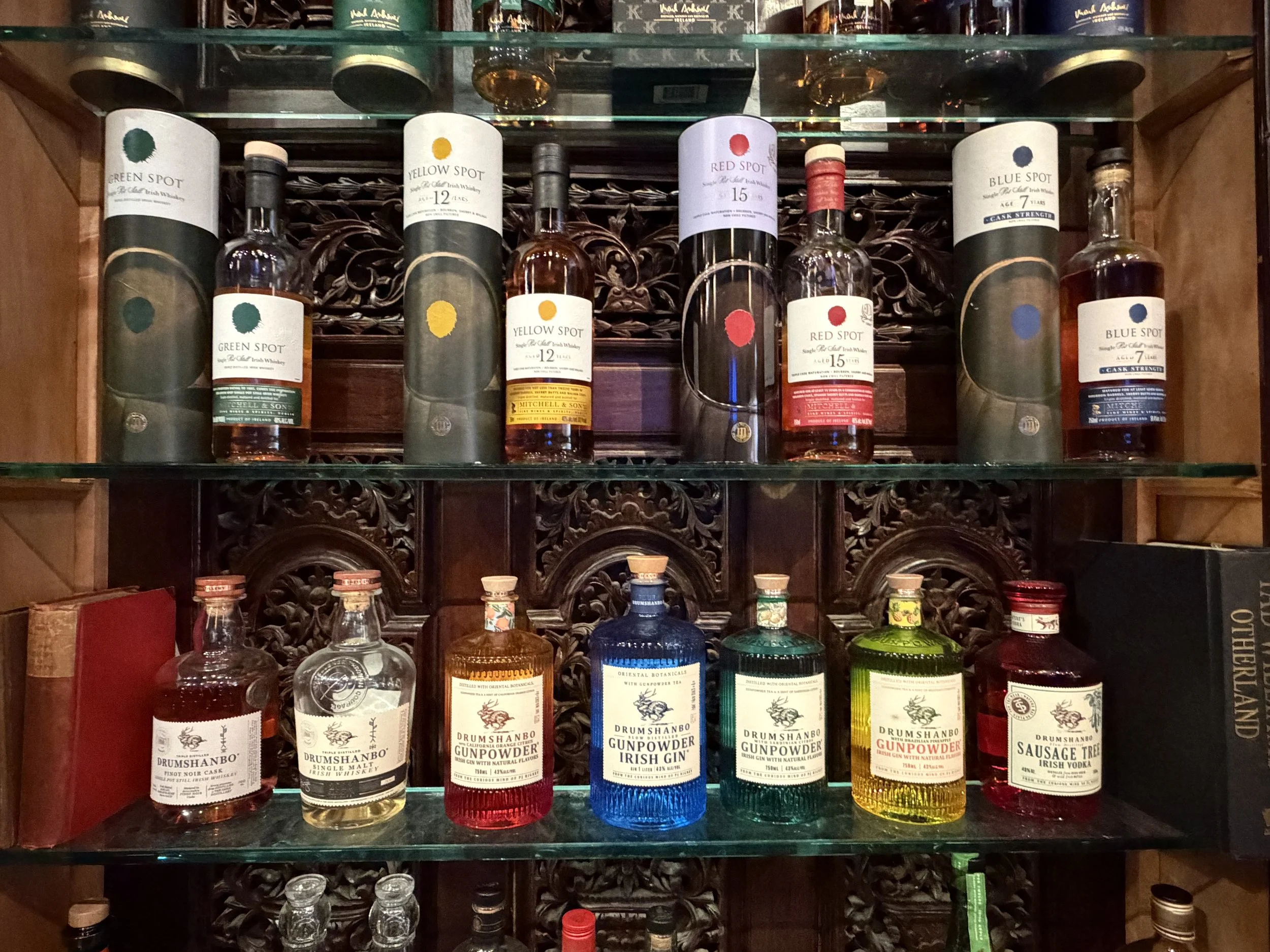 A display shelf with bottles of whiskey and gin, including Green Spot, Yellow Spot, Red Spot, Blue Spot, and Drumshanbo Gunpowder Irish gin and whiskey, arranged on glass shelves against a carved wooden backdrop.