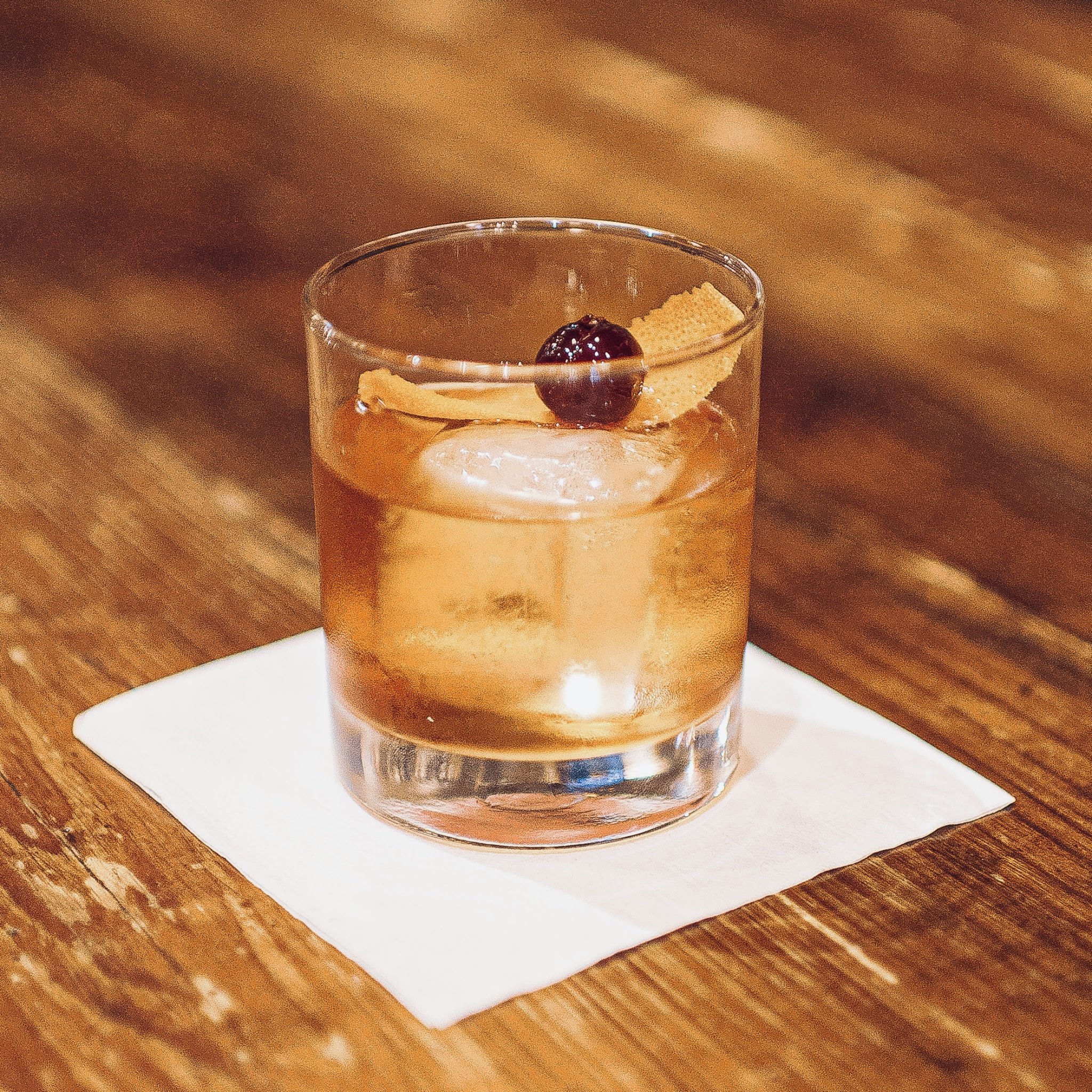 A glass of amber-colored cocktail with ice, garnished with a cherry, a lemon twist, and a label, placed on a white napkin on a wooden surface.
